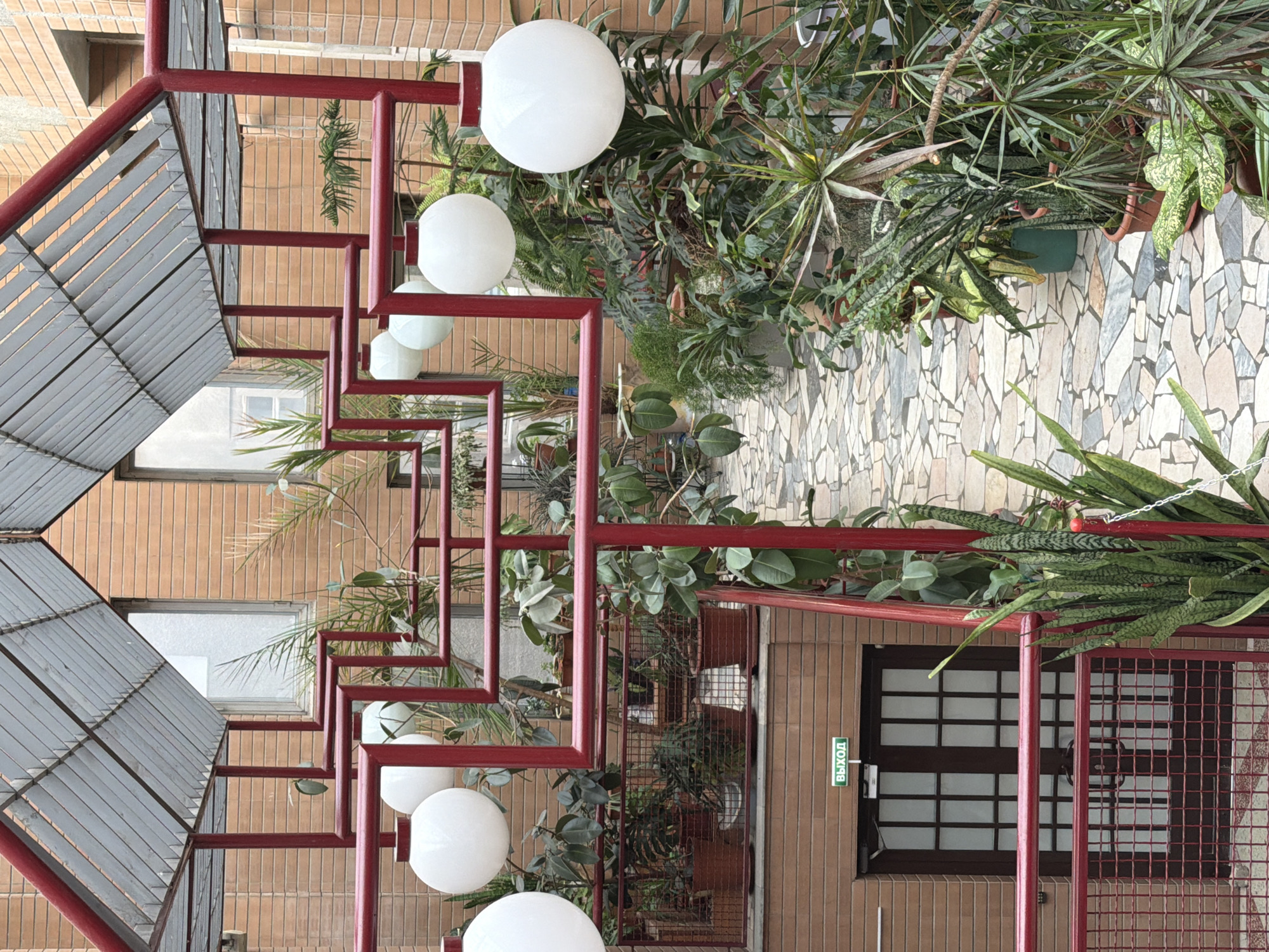 The image depicts an architectural structure adorned with vegetation and illuminated by spherical lamps.
The primary structure consists of a series of red metal frames, resembling a staircase or a canopy. These frames are positioned against a brick building, where windows are visible. Attached to these frames are large, white spherical lamps. Various potted plants and greenery cascade from the structure and the building, adding a natural element to the scene.
To the right, a mosaic-tiled walkway or wall is visible. In the lower part of the image, there is a door with a sign in Russian script, indicating "Exit." The setting is in Naberezhnye Chelny, Russia.