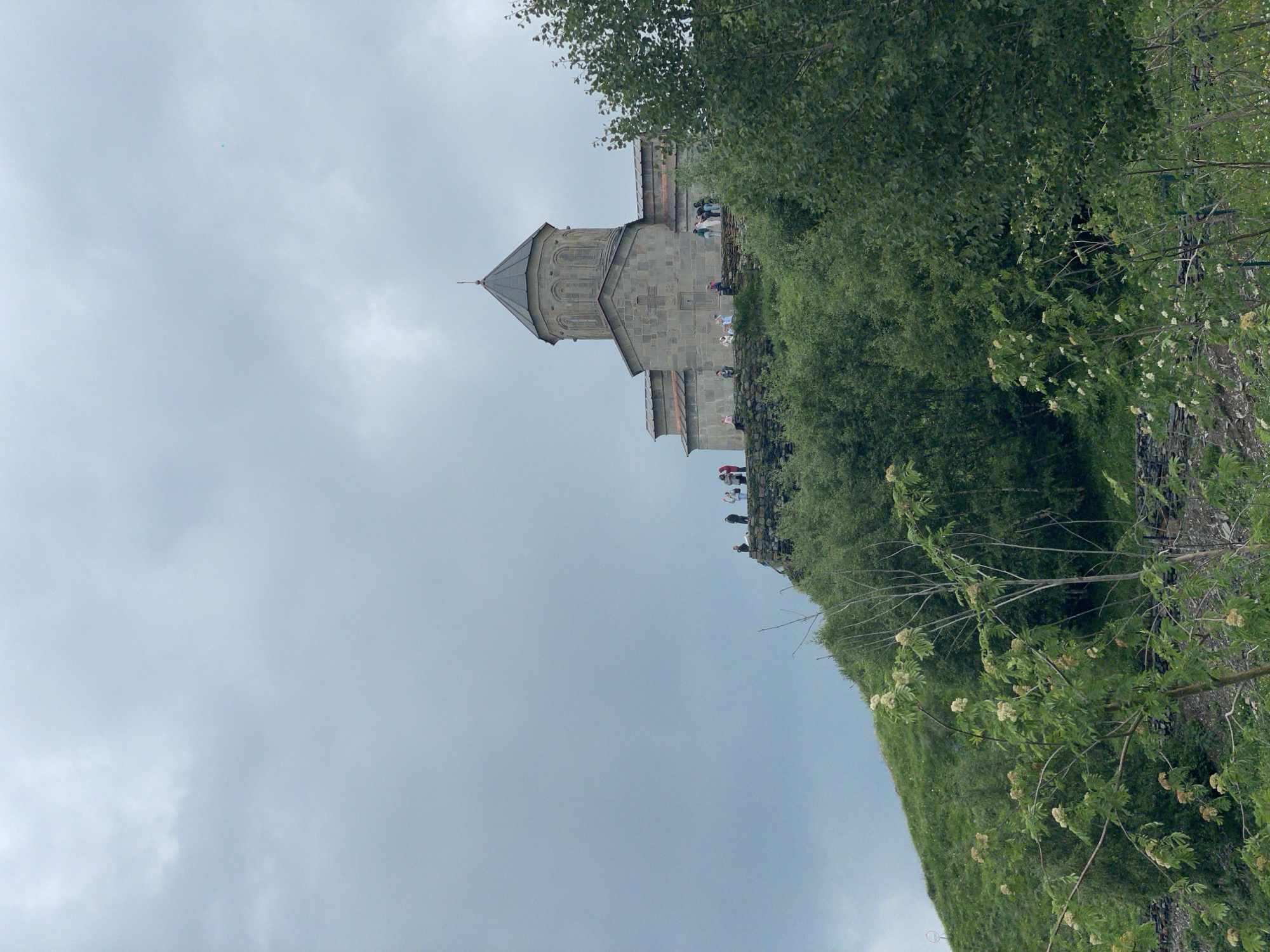 An exterior, eye-level, vertical shot features a stone church with a conical roof situated on a grassy hilltop in Stepantsminda, Georgia. The sky above is overcast and gray. The church has a cross relief above its entrance, and several people are visible on the stone terrace in front of it, appearing to be tourists. The foreground is filled with lush green trees and foliage, with some small white flowers on the branches of a tree in the lower left corner. A stone wall runs along the base of the hill where the church is located.