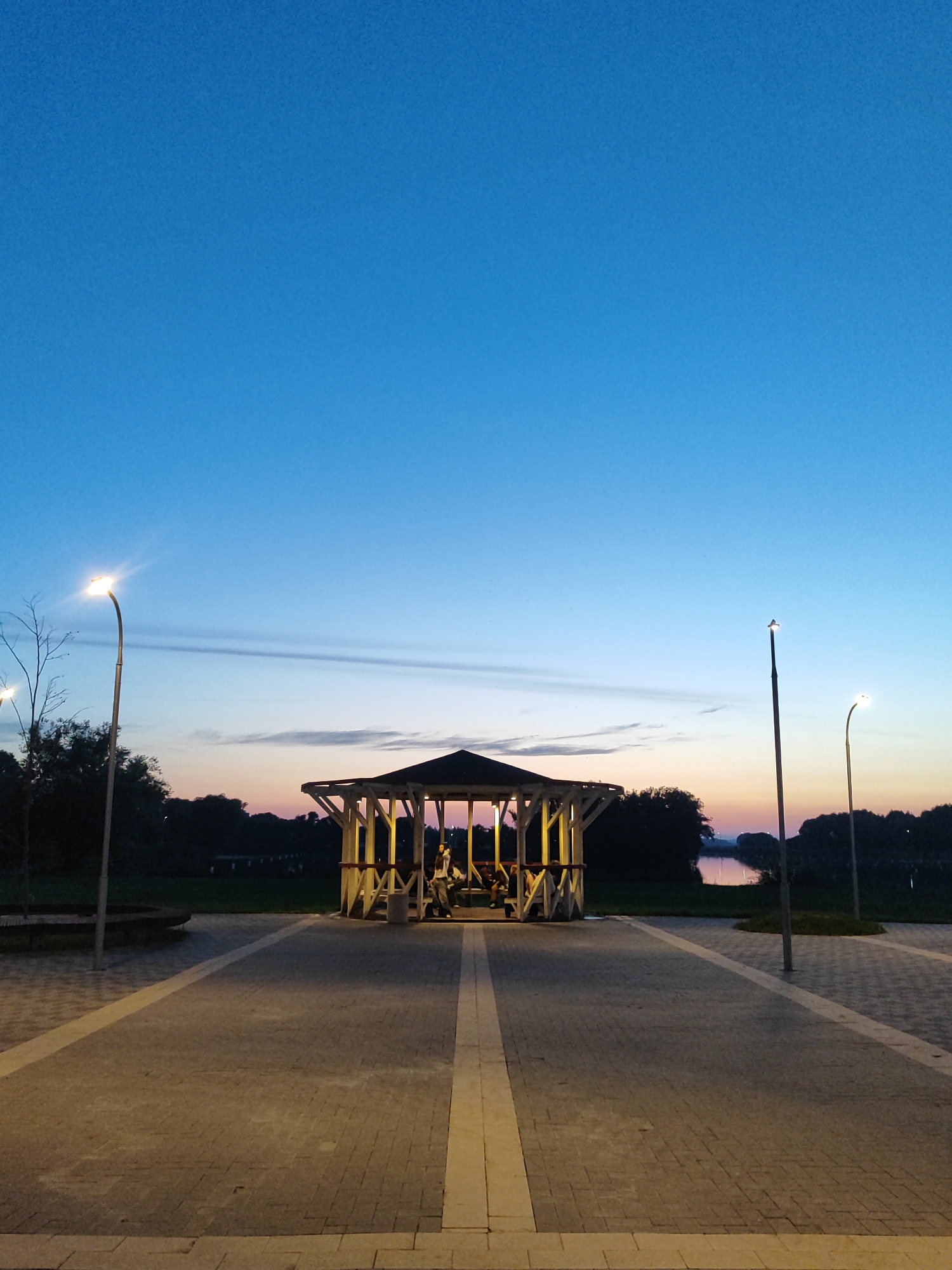 The image depicts a public park area in Bronnitsy, Russia, during the twilight hours. A wooden gazebo with white latticework stands centrally, illuminated by interior lights, and contains several individuals seated within. The ground is paved with interlocking stones, forming pathways and plazas. Two streetlights are visible, one on the left and one on the right, casting light onto the paved areas. Beyond the gazebo, a line of dark trees borders a body of water, reflecting the soft colors of the sunset and the sky, which transitions from a deep blue overhead to lighter hues near the horizon. The overall ambiance is tranquil and serene, suggesting a peaceful evening scene.