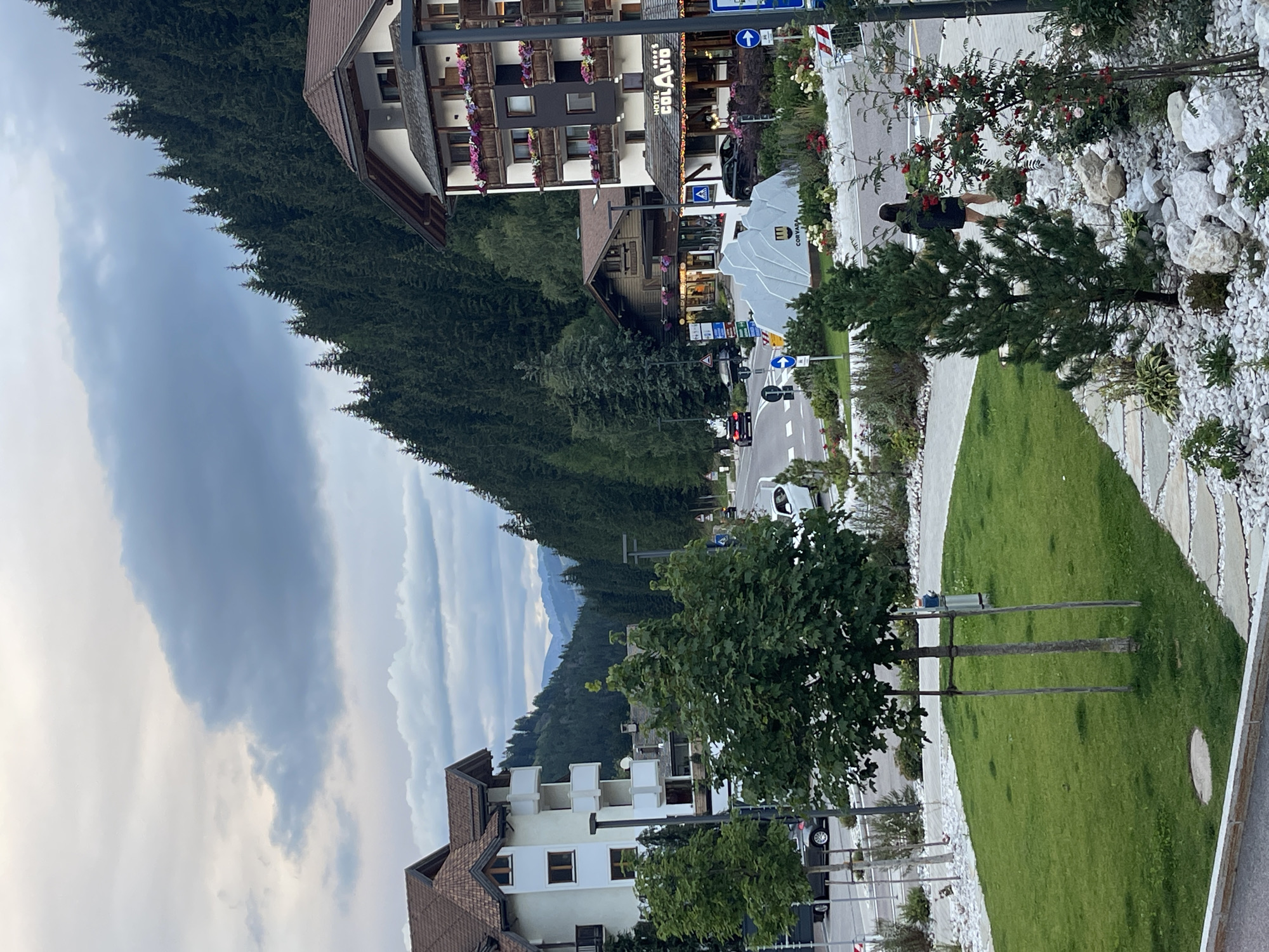 The image shows a street scene in Colfosco, Italy, with a hotel and other buildings lining the road. A woman in a black outfit is walking on the sidewalk, looking at her phone. In the foreground, there is a patch of grass with trees and decorative rocks. In the background, a dense forest covers a mountain slope. The sky is cloudy, and the overall impression is of a tranquil, natural setting.