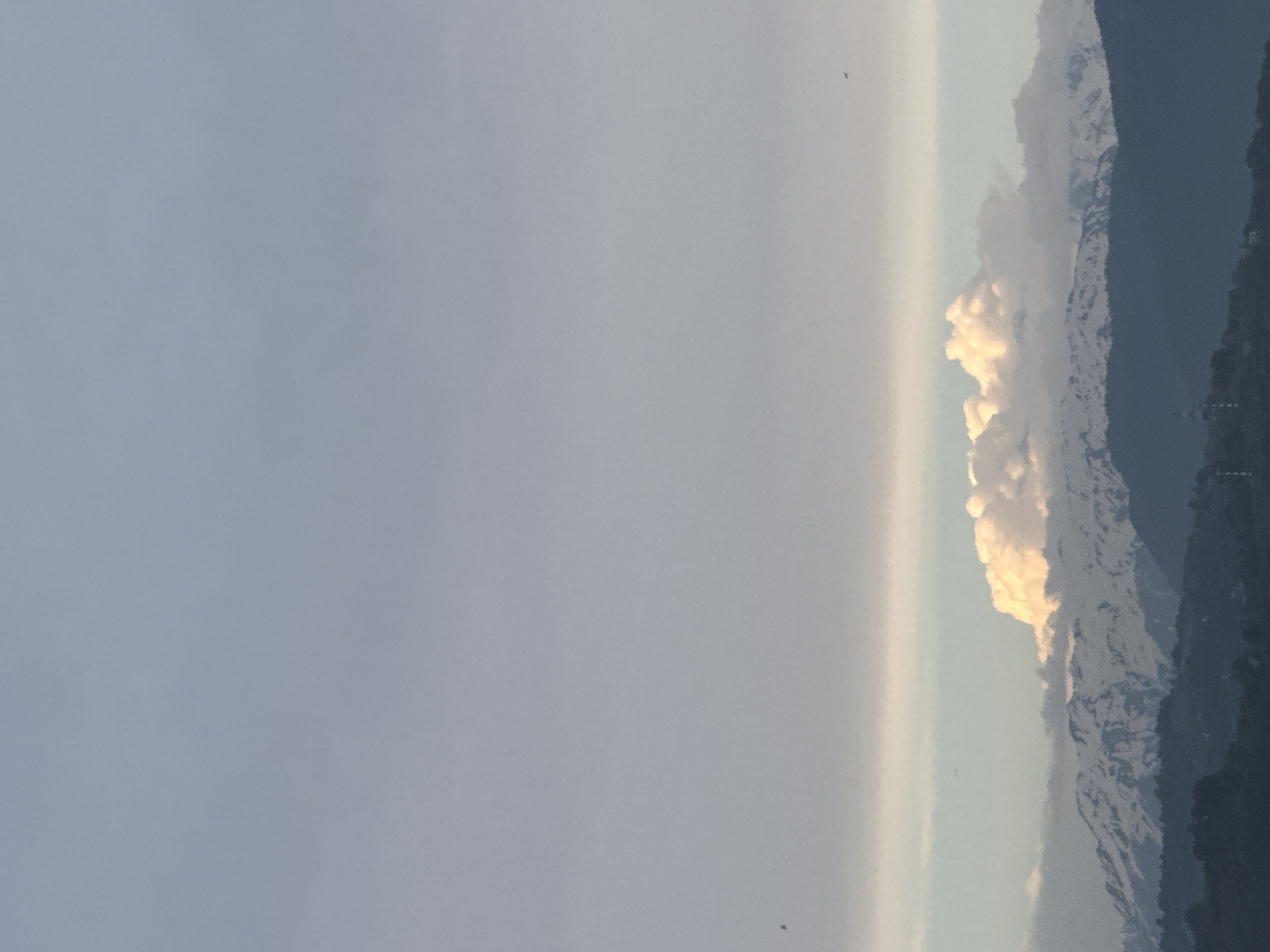 The image shows a cloudy sky with a line of mountains along the horizon. The mountains appear partially covered in snow. To the left of the mountain range is a large area of overcast sky. Two small specks, possibly birds, are visible in the sky. Given the prompt, this scene is likely located near Sochi, Russia.