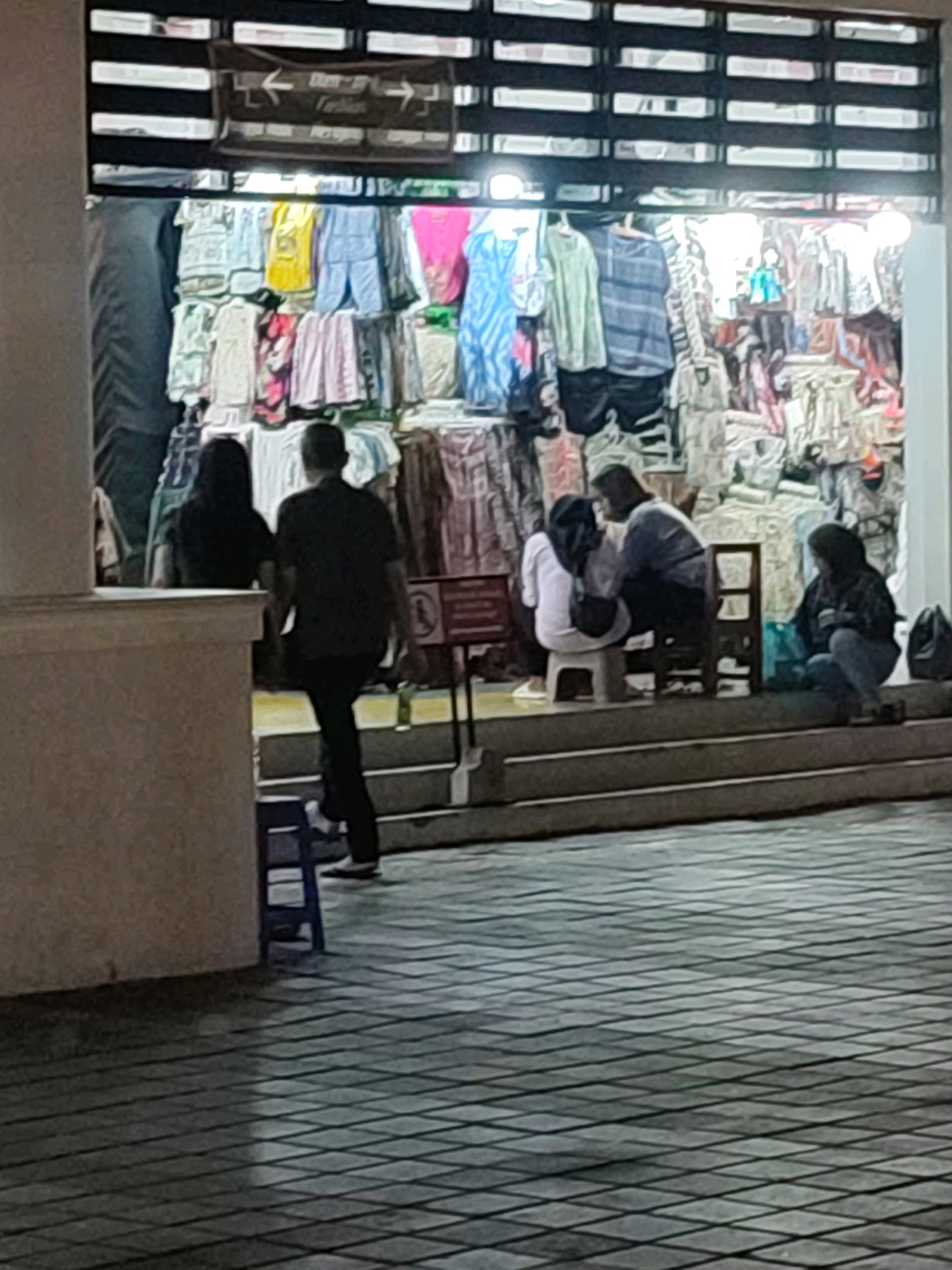 The image depicts a street-level view of a shop selling clothing, possibly located in the Malioboro area of Yogyakarta, Indonesia. The shop front is open, revealing racks of clothes, including shirts, pants, and dresses, in various colors and patterns. The lighting suggests it is evening or night.
In the foreground, there are several people. On the left, a man in a dark shirt and pants is walking away from the viewer, towards the shop. Next to him, a woman in a black head covering and black attire is standing, facing the shop. To the right, in front of the shop, three individuals are seated on steps or low stools. One person in a white top and black pants is seated on a white stool, facing away. Another person, possibly in a light blue shirt and dark pants, is seated behind them, facing them. A third person in a dark jacket and blue jeans is seated on the steps to the right, looking towards the shop. A dark bag is visible next to this person.
The ground in front of the shop and extending into the foreground is paved with a diamond-patterned tile. Steps lead up to the shop entrance. A blue plastic stool is placed on the tiled area in the lower left quadrant of the image. In the background, above the shop entrance, a sign with arrows pointing left and right is visible, possibly indicating directions within a market or area.