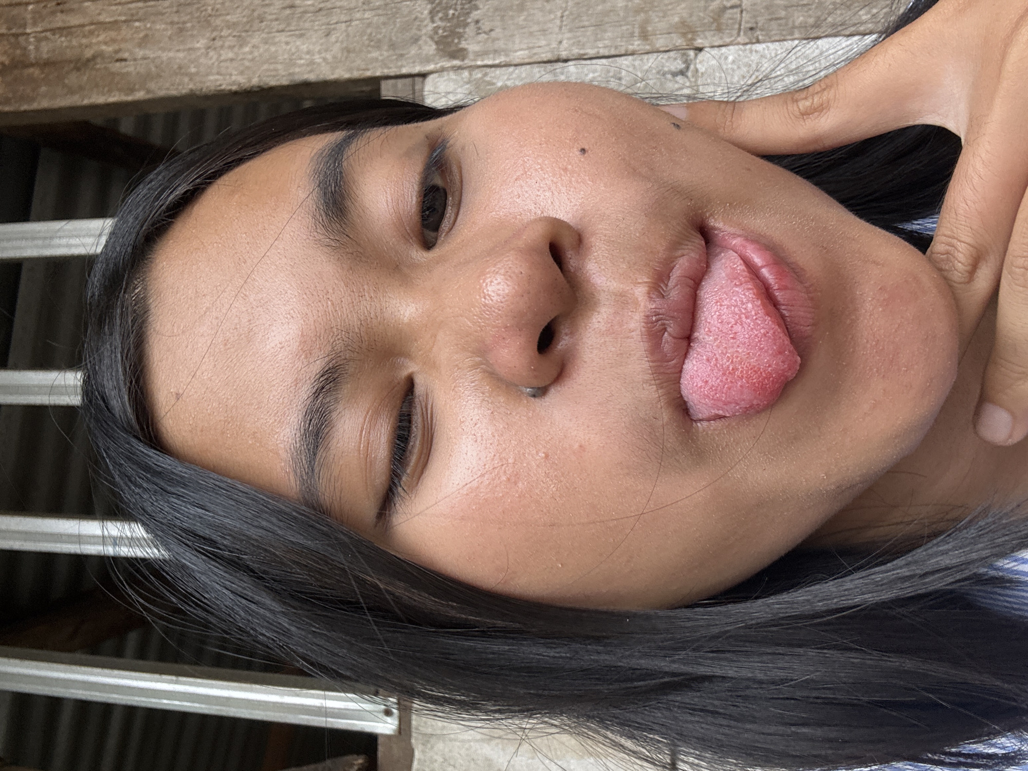 A close-up, waist-up, outdoor selfie shows a young woman with long, dark hair winking and sticking her tongue out. She is wearing a blue and white striped shirt. Her face is slightly tilted, and her right hand is touching her cheek. The background consists of a wooden fence and metal bars, suggesting a rustic or possibly unfinished structure. The lighting is natural, indicating it is daytime. The location is East Jakarta, Indonesia.