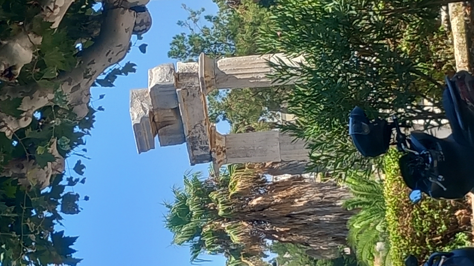 The image captures an ancient Greek ruin consisting of two large fluted columns topped with an entablature. The structure is partially obscured by lush green foliage in the foreground and the canopy of a large tree with mottled bark at the top. In the background, a tall palm tree with dry, drooping fronds stands to the left of the ruins, and other trees with green foliage are visible behind and to the right. The sky is a clear, bright blue. In the lower right of the frame, the handlebars and seat of a dark-colored scooter are visible, with a dark helmet resting on the seat, suggesting the presence of a parked vehicle. The scene appears to be outdoors, possibly in a park or historical site. This ruin is likely part of the Agora of Kos, the ancient marketplace of Kos Town.