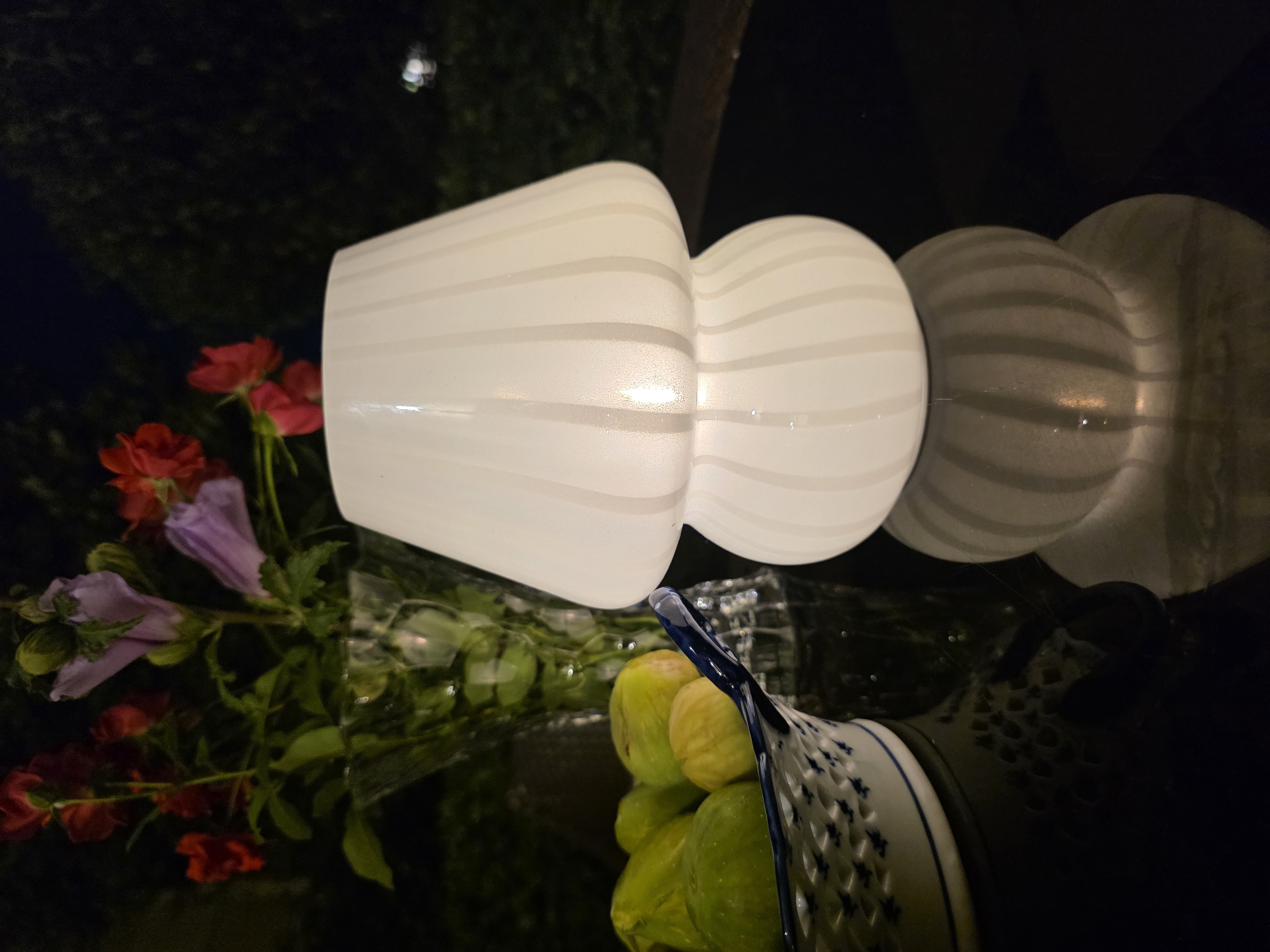 An overhead, medium shot captures a still life arrangement on a dark, reflective surface, likely a table, under low-light conditions at night. In the foreground, a white, mushroom-shaped lamp with vertical ribbing emits a warm glow from within. To the left, a white, oval bowl with blue floral accents contains a cluster of green figs. Behind the figs, a clear, ornate vase holds a bouquet of red and purple flowers. The background is a dark, out-of-focus expanse of greenery and a hint of distant, blurred lights. The scene is set in an unknown outdoor location, possibly a patio or garden, in Unknown, Unknown. There are no people or animals visible.