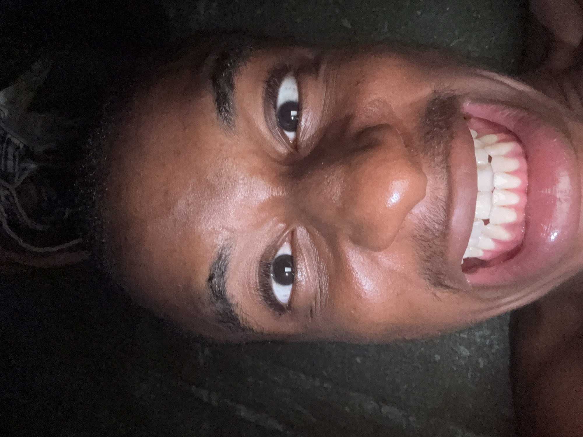 A close-up, upward-facing selfie shows a Black individual's face against a dark, textured background that may be a wall or ground. The individual's eyes are wide open, and they are smiling broadly, revealing a full set of white teeth and pink gums. The skin appears moist and reflects light. The individual's hair, styled in dreadlocks, is visible at the top of the frame. The scene is dimly lit. This image was taken at Nkpor, Nigeria.