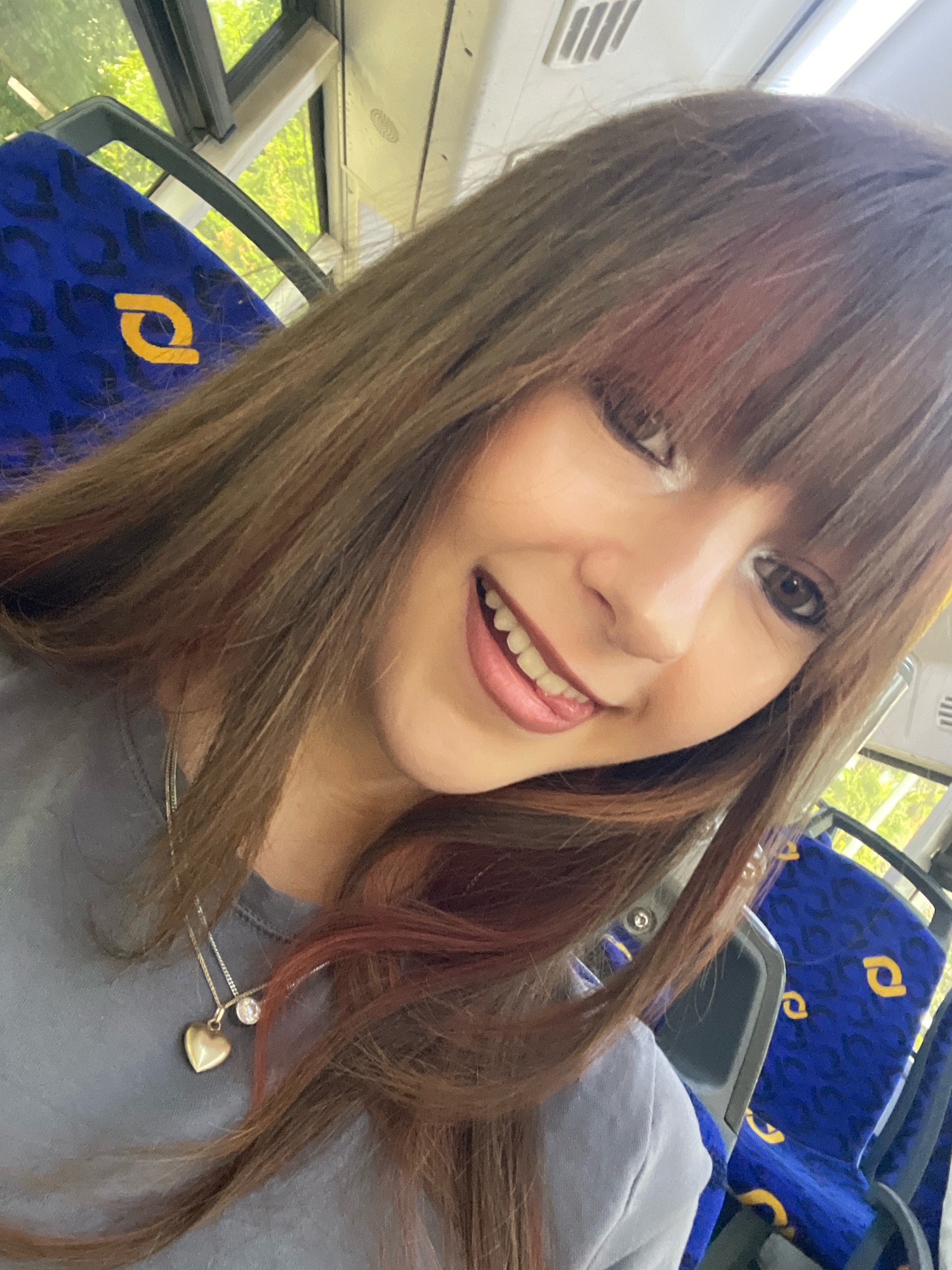 The image shows a close-up, selfie-style shot of a young woman with long, brown hair, some of which has reddish highlights, sitting on a bus. She is smiling directly at the camera, with her tongue slightly visible. She is wearing a gray t-shirt and a silver necklace with a heart-shaped pendant and a clear stone. The background of the image shows blue bus seats with a yellow logo, and through the windows, greenery is visible, suggesting the bus is in a location with trees. The lighting appears to be natural sunlight, indicating it is daytime. The scene is likely on a public transport bus within the city of Raanana, Israel, given the context.
