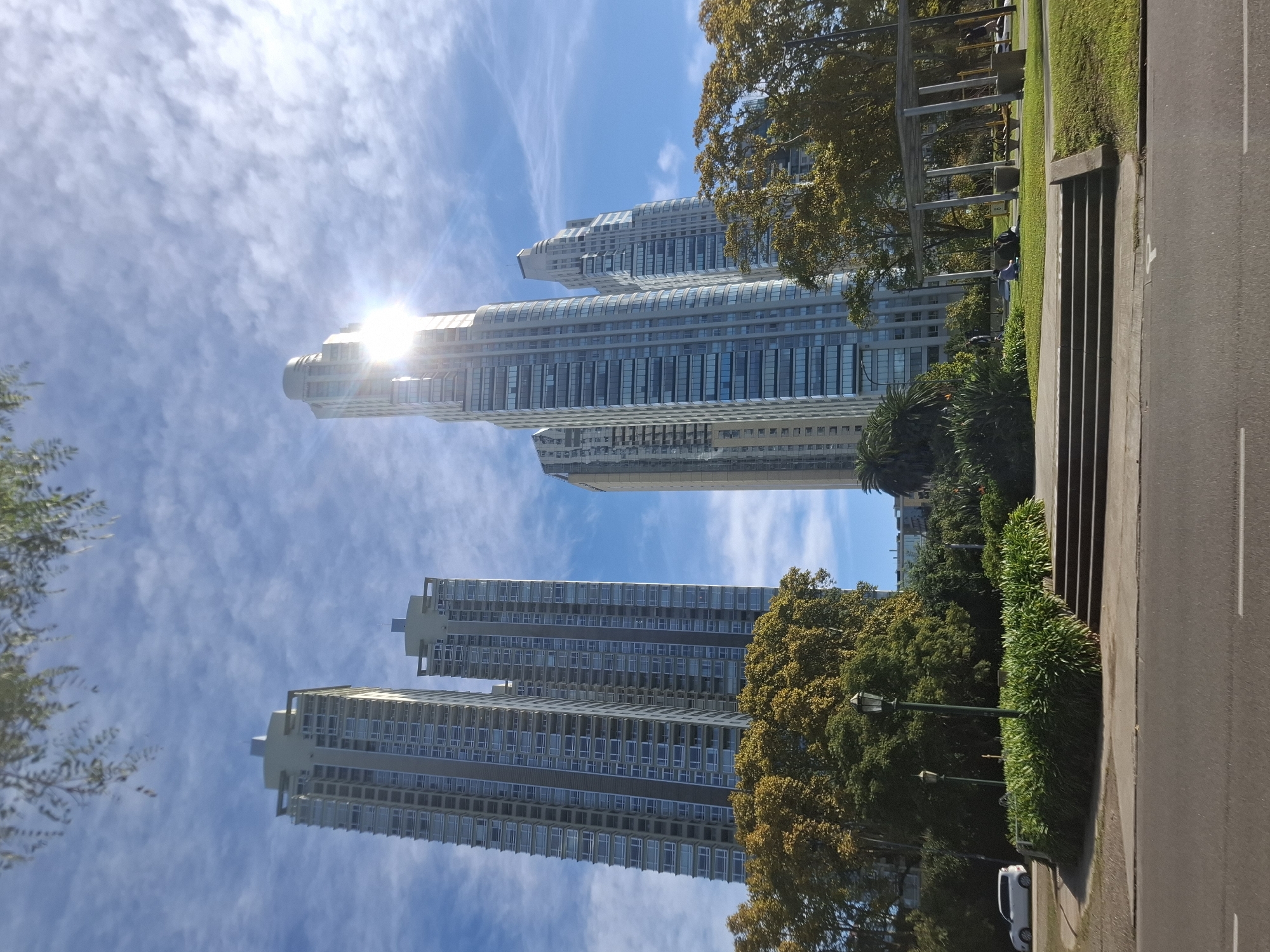 The image depicts a view of the River View towers in the Puerto Madero neighborhood of Buenos Aires, Argentina. The perspective is from street level, looking up at the buildings against a bright blue sky with wispy clouds. The sun is reflecting off the upper portion of one of the towers, creating a bright glare. To the right of the towers, there is a grassy area with trees, bushes, and streetlights. Adjacent to this is a set of concrete stairs leading up to a higher level of the park. A portion of a road is visible on the extreme right side of the frame. A white car is parked near the road. The composition suggests an upward angle and urban setting with a mix of modern architecture and natural elements.