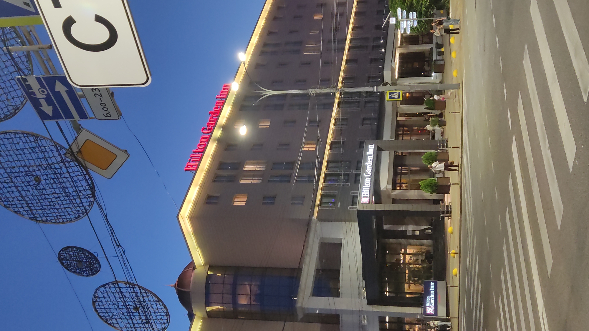 The image depicts a street scene at dusk in Krasnodar, Russia, with a Hilton Garden Inn hotel as the prominent subject. The hotel is a large, multi-story building with its illuminated "Hilton Garden Inn" signage clearly visible in bright red letters. The building's facade features many windows, some of which are lit from within, suggesting occupants or activity inside.
In the foreground, a pedestrian crossing (zebra crossing) is visible on the asphalt road, indicating an urban environment. Several people are present in the scene, with some walking on the sidewalk near the hotel entrance and others possibly seated at outdoor dining areas. The lighting suggests it is early evening or dusk, with streetlights and the hotel's external lighting casting a warm glow. The sky is a deep blue, indicating the transition from daylight to night.
Several street signs are visible, including a circular sign with a black "C" and a rectangular sign with "6.00-23", likely indicating operating hours or time restrictions. There are also decorative wireframe circular objects suspended from wires, adding an artistic element to the urban landscape. The overall atmosphere is that of a typical city evening, with the hotel being a focal point of activity.