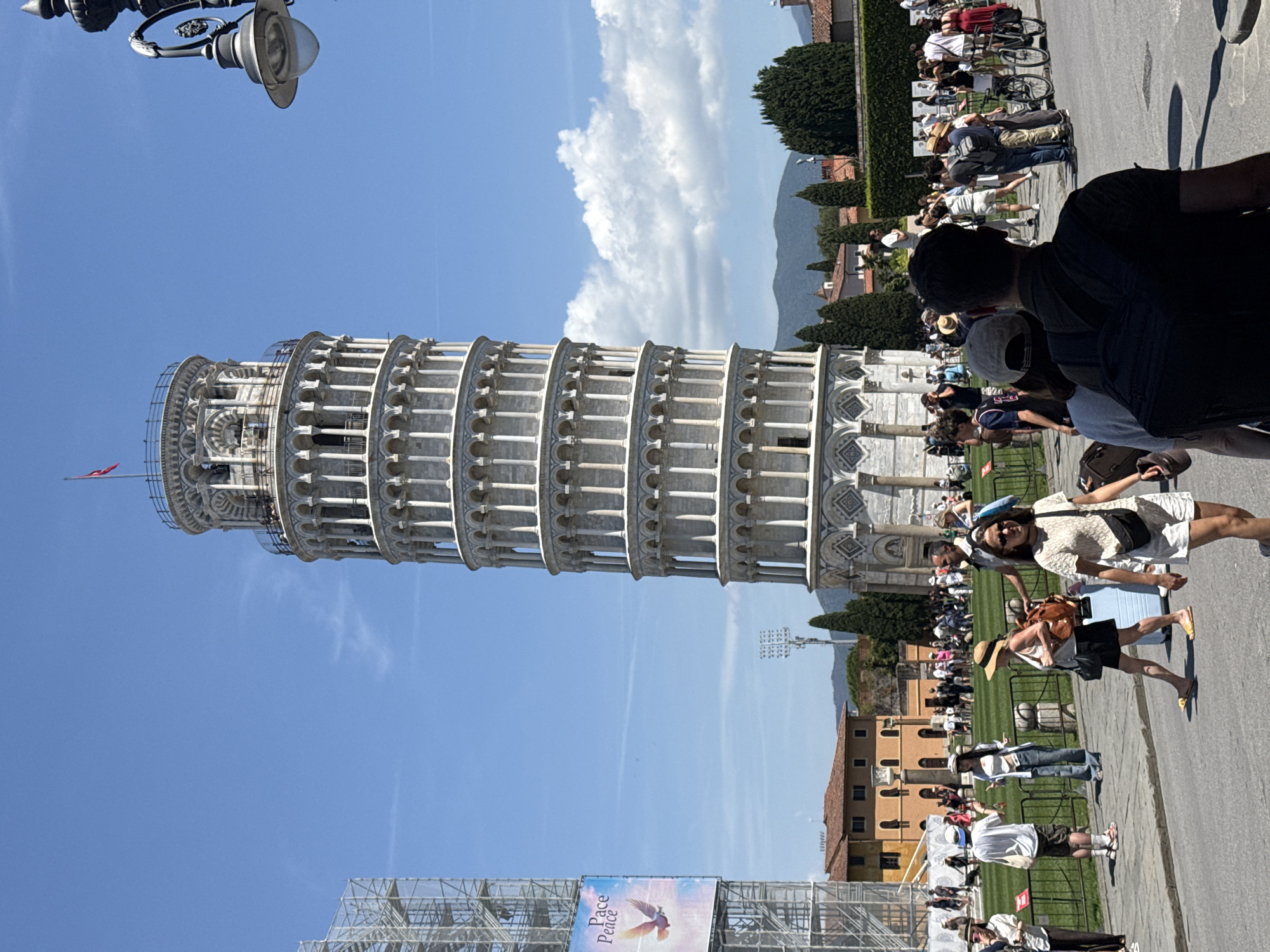 The image depicts the Leaning Tower of Pisa in Pisa, Italy, under a clear blue sky with scattered clouds. The tower, a cylindrical bell tower made of white marble with arcades and columns, dominates the center of the frame. The lower portion of the tower shows some decorative geometric patterns. Scaffolding is visible on a building to the left of the tower, and a banner with text and an image of a bird is attached to it.
In the foreground and middle ground, a crowd of tourists are visible on a paved area and a grassy lawn. People are walking, standing, and observing the tower. Some individuals are dressed in casual summer attire, including hats, shorts, and t-shirts. There are a few bicycles parked in the background. The environment appears to be a public space, possibly a piazza, with some trees and buildings in the background. The overall scene is one of a sunny day with many visitors enjoying the famous landmark.
