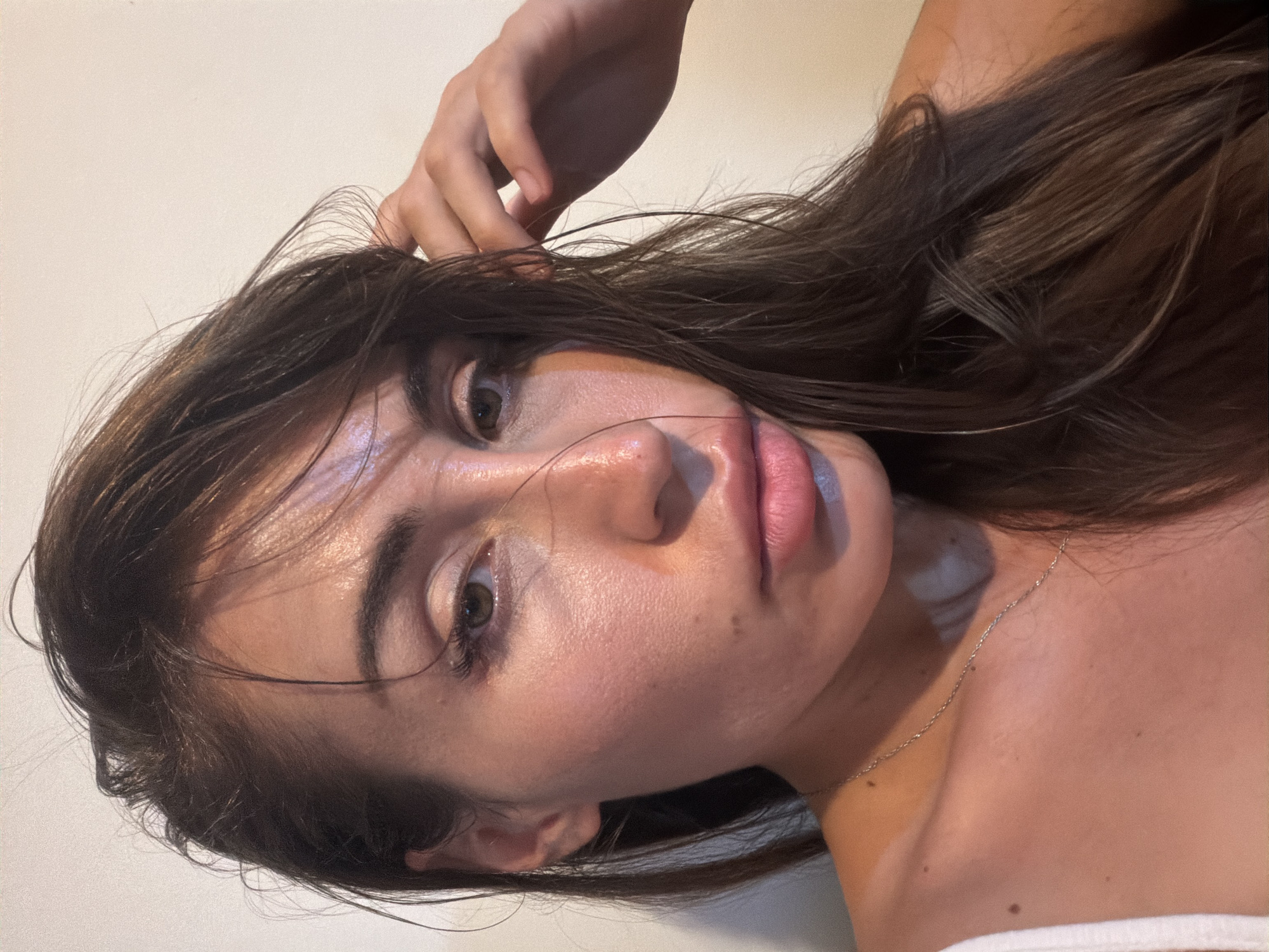 The image shows a close-up, head-and-shoulders shot of a woman with long, dark brown hair and fair skin. She is looking towards the camera with a neutral expression. Her hair is slightly messy, with some strands falling across her face and shoulder. Her left hand is raised, touching her hair near her temple. She is wearing a white strap, likely from a tank top or bra, and a delicate silver chain necklace. The background is a plain, off-white wall. The lighting appears to be artificial and warm, casting highlights on her face and neck, suggesting the photo was taken indoors. No specific location in Tersefanou, Cyprus is identifiable from the image.