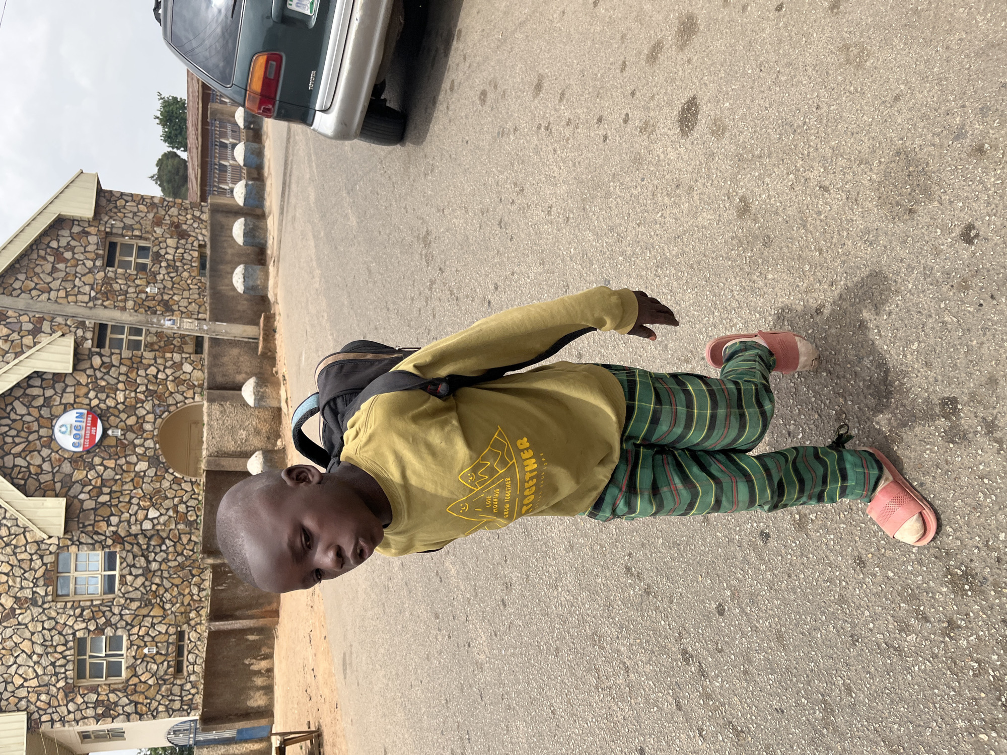 A young boy is standing on a paved street. He has a shaved head and is wearing a yellow long-sleeved shirt, green plaid pants, and pink sandals. He is wearing a backpack. He is standing next to a building of stonework that is of COGIN school in the city of Jos, Nigeria. A parked vehicle is visible in the background.