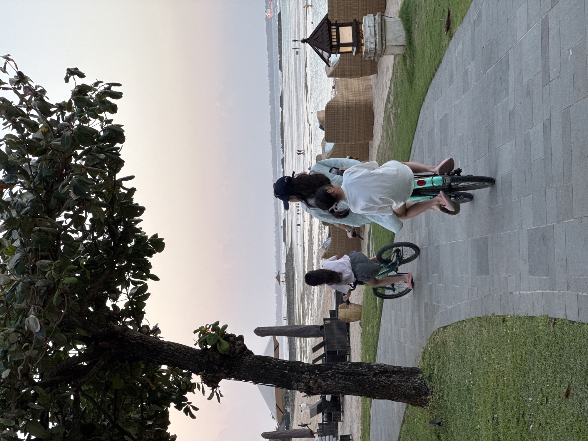 This image shows a paved pathway winding through a grassy area next to a beach. The path is bordered by green grass on the left and a beach with lounge chairs and umbrellas on the right. In the background, the ocean stretches to the horizon, with a few people visible on the shore. The sky is a soft gradient of pink and orange, suggesting either sunrise or sunset.
Three individuals are riding bicycles on the pathway. The foremost rider is a child wearing a white t-shirt and pink flip-flops, riding a teal bicycle. Behind them, another child on a bicycle faces away from the camera. To the left, a third child with long dark hair is also cycling. All three individuals appear to be enjoying a leisurely ride in this tropical setting. The overall scene evokes a sense of relaxation and family activity in Kuta Selatan, Indonesia.