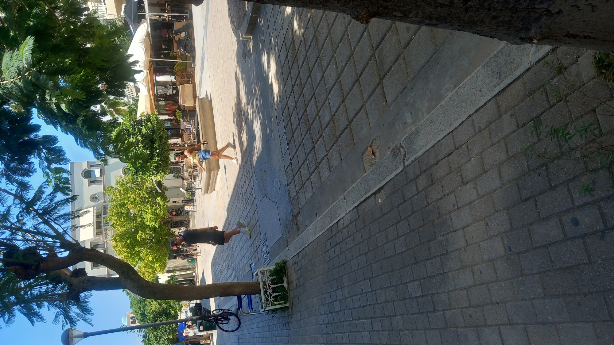 The image depicts a street scene in the city of Kos, Greece, likely during the daytime. In the foreground, a cobblestone sidewalk is visible, with a raised curb running along its edge. Several trees provide shade, with their leaves casting dappled shadows on the ground.
Two individuals are walking in the middle ground. A person wearing a dark t-shirt and shorts is walking to the left, while a person in a light-colored top and denim shorts is walking to the right. In the background, there are buildings with white facades and awnings, suggesting commercial establishments such as cafes or restaurants. A fountain is also visible, and tables and chairs are set up outside. A bicycle is parked on the left side of the image, near a street lamp. The overall environment suggests a bustling urban area with outdoor seating for patrons. The sky is clear and blue.