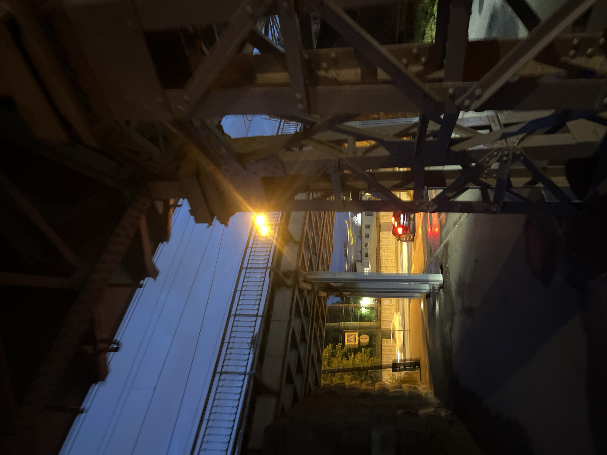 The image is a low-angle, nighttime photograph taken from beneath an elevated railway bridge in Saint Petersburg, Russia. The perspective is dominated by the robust, dark metal support structure of the bridge, with its rivets and beams clearly visible. Beyond this immediate foreground, the railway track itself is visible, lined with a metal railing and illuminated by a bright yellow lamp.
Below the railway bridge, a road is visible, wet from recent rain, with reflections of lights on its surface. A single vehicle, a dark-colored car with its red taillights illuminated, is driving away from the camera. To the left of the road, there is a pedestrian crossing sign with a yellow triangular warning symbol and a white pedestrian silhouette. Further down the road, indistinct buildings can be seen in the background, illuminated by streetlights. The sky above is a deep twilight blue. The overall atmosphere is urban and industrial, with a sense of stillness due to the nighttime setting.