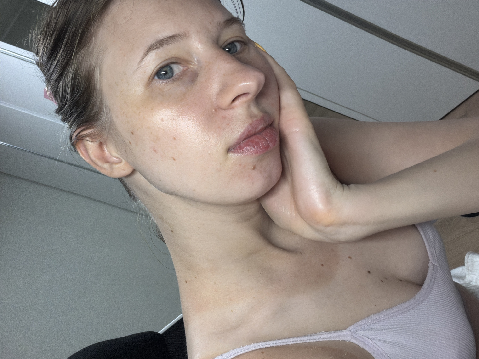 A young woman is captured in a close-up selfie, her head tilted to the side with her right hand propping up her chin. She is wearing a light purple tank top. Her skin is fair with visible freckles and some blemishes. Her eyes are blue and her lips are slightly parted. The background appears to be an indoor setting with a grey wall and a white door or cabinet. The lighting suggests an interior environment. The image is shot from a high angle. The woman's hair is brown and pulled back, with some strands falling loose. The overall impression is a casual, unposed moment.