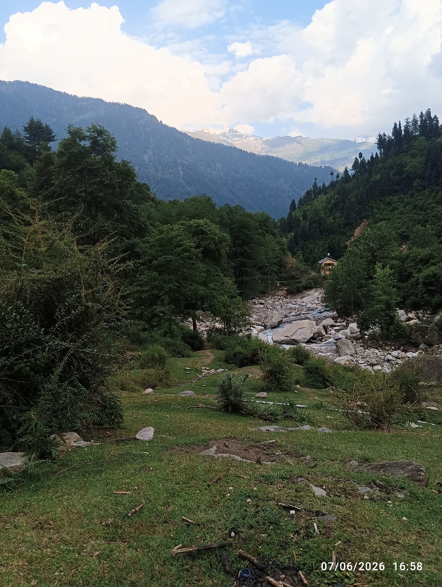 The image depicts a mountainous landscape in Old Manali, India, under a partly cloudy sky. Dominant features include densely forested mountainsides flanking a central valley. In the far distance, snow-capped peaks are visible beneath a horizon of large white clouds against a pale blue sky.
The midground is dominated by a rocky riverbed through which a light blue-green stream flows from the background towards the foreground. Numerous light-colored boulders and smaller rocks are scattered throughout the river and along its banks. On the right side of the river, situated on a vegetated slope, a small, light-yellow building with a dark roof is visible, with what appears to be utility lines extending across the river towards it. Several small human figures are discernible in and around the riverbed near this building, appearing to be engaged in leisure activities such as wading or sitting.
The immediate foreground consists of a grassy area interspersed with bare earth and scattered rocks. Various green bushes and smaller trees, including a dense, thorny shrub on the left, populate this space. Fallen branches and some scattered debris, including a blue plastic cap, are visible on the ground. A faint dirt path appears to lead from the foreground towards the river. The overall environment is one of natural rugged terrain with evidence of human presence. A date and time stamp "07/06/2026 16:58" is present in the bottom right corner of the image.