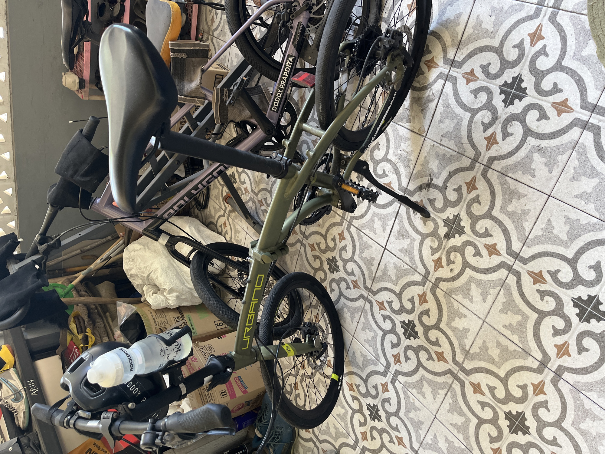 This image depicts a close-up, eye-level view of two folding bicycles parked indoors, likely in a storage area or garage, on a patterned tile floor. The foreground bicycle is a green folding bike with the brand name "URBANO" in yellow lettering. It has a black saddle, black handlebars with grey grips, and a water bottle cage mounted to the frame holding a clear water bottle with a "ROCKBROS" logo. The front wheel of this bicycle is turned towards the left. Behind it, a second folding bicycle with a purple frame and black components is partially visible. The rear wheel of the green bicycle shows a derailleur and disc brake. The environment includes various storage items and boxes in the background. The tile floor features a repeating ornate pattern with grey, white, and brown elements. There is no indication of specific location in Sidoarjo, Indonesia, beyond the general context of an indoor setting.