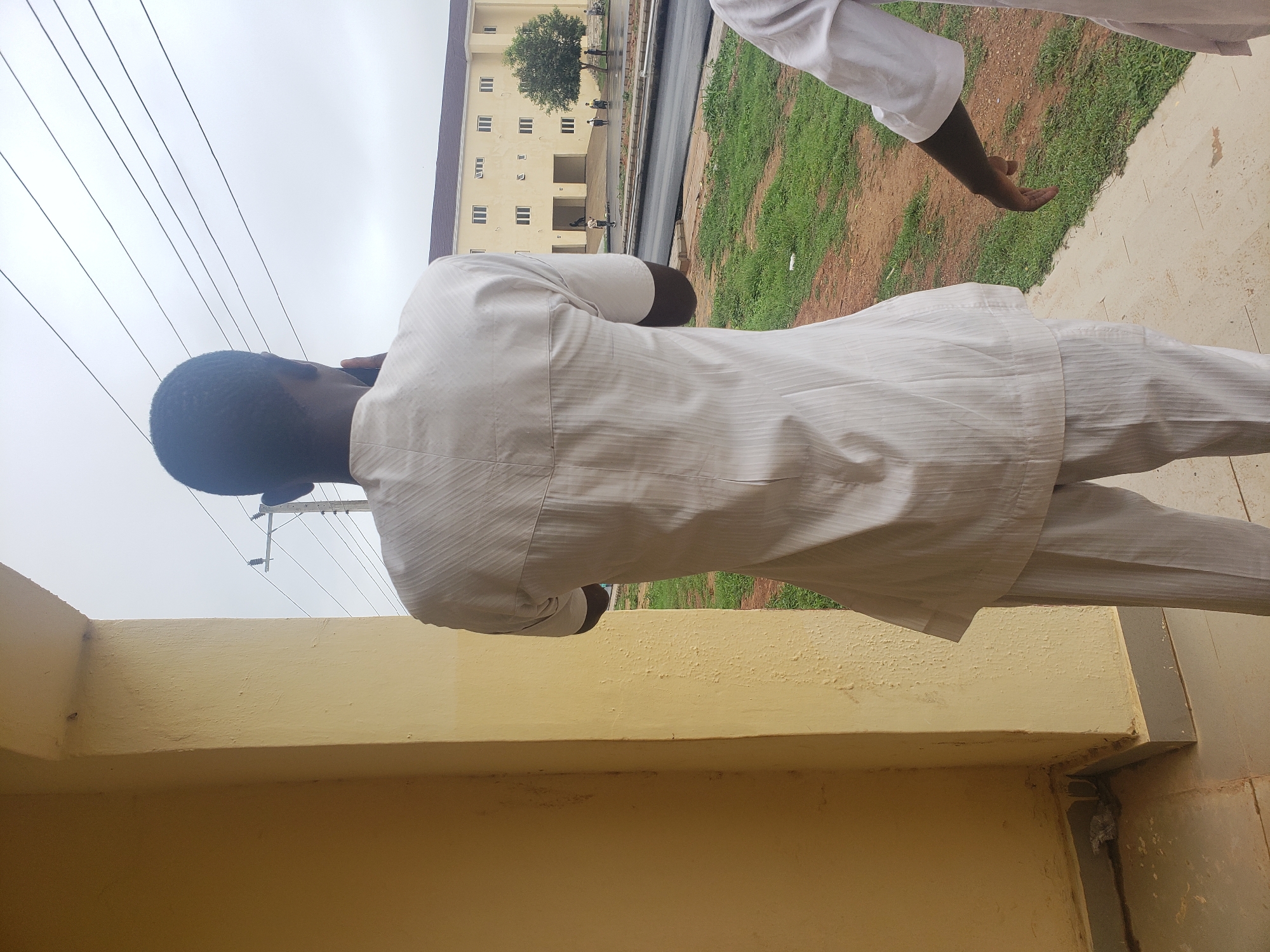 The image depicts a street scene in Gumel, Nigeria, with a focus on the rear view of two individuals dressed in white traditional attire. The foreground features a textured yellow wall and a concrete floor. In the mid-ground, a paved road is visible, with patches of green grass and red soil on either side. In the background, a multi-story building with a tiled roof stands, and several trees are planted in front of it. Power lines stretch across the overcast sky. One person is facing away from the camera, possibly engaged in a conversation on their phone, while another person's arm is visible on the right side of the frame, extended towards the street. The scene suggests a casual outdoor setting within the city.
