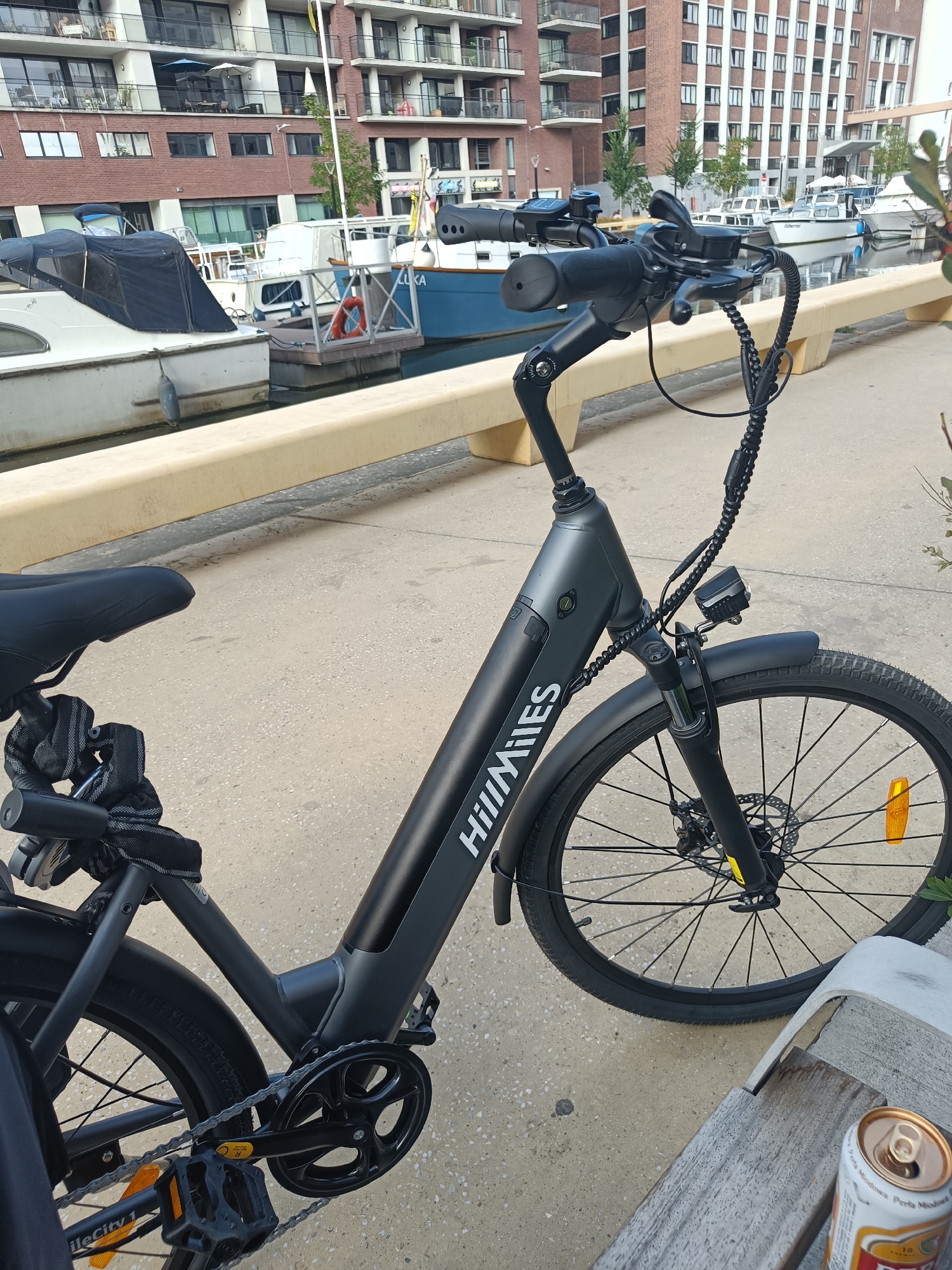 The image shows an electric bicycle parked on a concrete path next to a canal. The bicycle is black and has "HillMiles" written on the frame. The handle bar is turned to the left, and the front wheel is facing towards the canal. In the background, there are several boats docked in the canal, and apartment buildings with red brick facades and white balconies. A can of beer is visible on a wooden bench in the foreground. The scene is set in an urban environment with a waterfront.