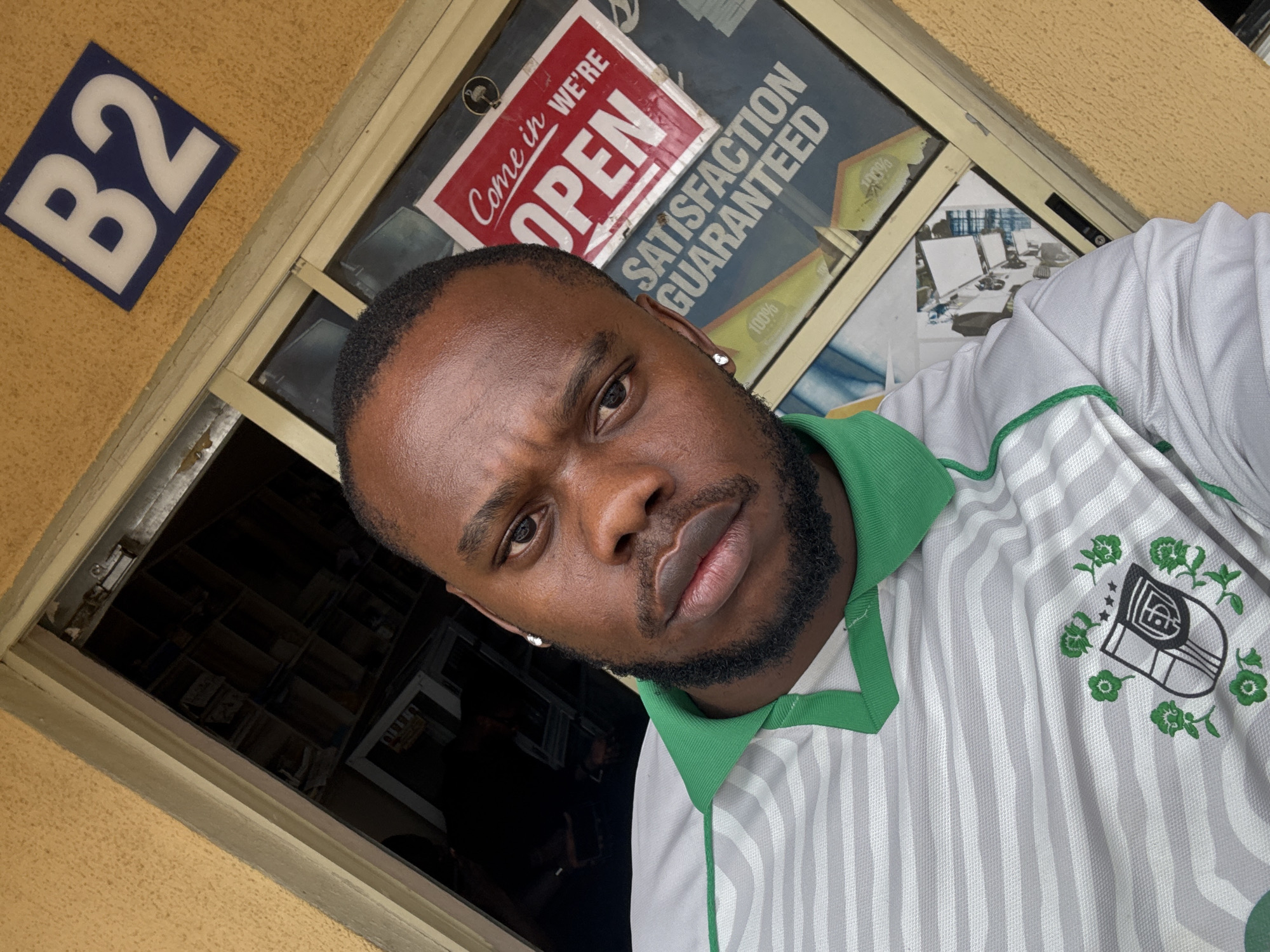 A young Black man is captured in a selfie, angled from slightly above and to his right, giving a close-up of his head and torso. He has a dark, well-groomed beard and short hair. He is wearing a white and grey striped polo shirt with a green collar and green trim, featuring an emblem on the right chest with green flowers, a shield-like logo, and stars. He wears a silver stud earring in his left ear. His expression is serious, with a slight frown and direct gaze towards the camera.
The background shows the exterior of a building and a window. To the upper left, a blue square plaque with white lettering reads "B2" against a textured, light brown or beige wall. Behind the man's head, through what appears to be a glass window or door, a red and white sign is visible, stating "Come in we're OPEN". Below this, another sign is partially visible with the text "SATISFACTION GUARANTEED" and "100%". The reflection in the glass also shows what looks like a computer setup inside, with multiple monitors. The interior seen through the window is dimly lit, suggesting it might be dark inside the building or the exterior light is reflecting strongly. The overall setting appears to be a commercial building or shop entrance in Lugbe, Nigeria, given the provided location context.