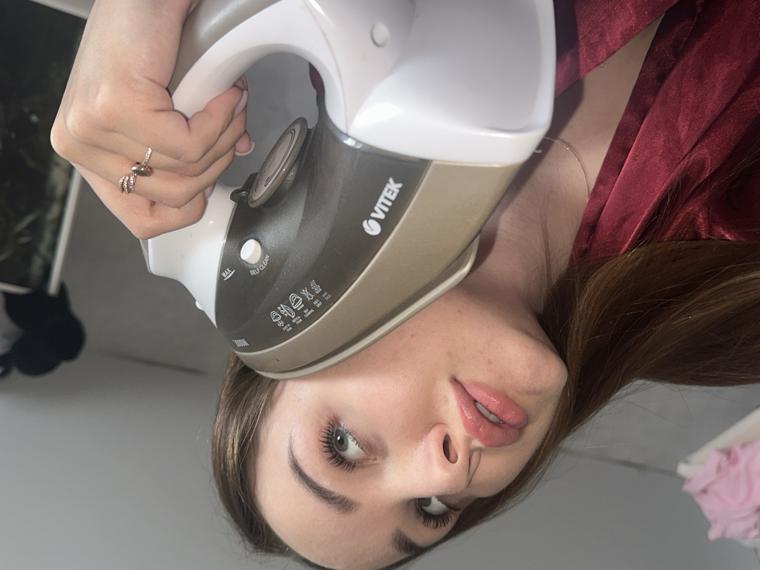 This is a close-up, rotated portrait of a young woman posing playfully indoors in Chaikovsky, Russia. The image is oriented with the woman's head to the left and her body extending upwards.
The woman, likely in her 20s or early 30s, has medium-brown hair, fair skin, and full, slightly parted lips with pink lip gloss. Her eyes are light-colored and accentuated with mascara. She wears a reddish-brown satin garment, possibly a robe, and a delicate chain necklace. A multi-banded ring adorns her right hand, which firmly grasps a white and gold/bronze VITEK brand steam iron.
The iron's soleplate is positioned humorously against her right temple or forehead. Visible text on the iron includes the brand name "VITEK," along with "MAX," "SELF-CLEAN," and several small icons indicating various fabric settings and functions like steam and spray.
The background is simple and out of focus, consisting mainly of a plain, light-colored wall. Blurry dark shapes and a hint of pink fabric are discernible in the periphery. The overall activity depicted is a staged, humorous pose rather than an actual use of the iron for its intended purpose. The scene suggests a lighthearted or quirky mood.