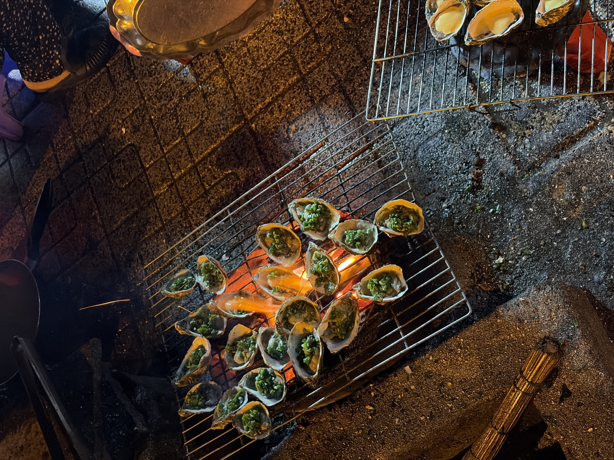 Oysters are being grilled over an open flame on a metal grate in Bac Nha Trang Ward, Vietnam. The oysters are topped with chopped green onions and appear to be cooking in their shells. The grilling is taking place outdoors at night, indicated by the artificial lighting and the dark surroundings. The ground is paved with textured tiles, and there are hints of charcoal and ash from the fire. In the background, there are glimpses of people's legs and feet, suggesting a street food setting. One person is wearing a dark, patterned fabric shoe with a yellow accent. To the right, a golden serving tray with an ornate rim is visible. A metal tong is positioned above a dark pot or wok in the upper left corner of the image. The overall scene depicts a casual, nighttime outdoor dining experience focused on grilled seafood.