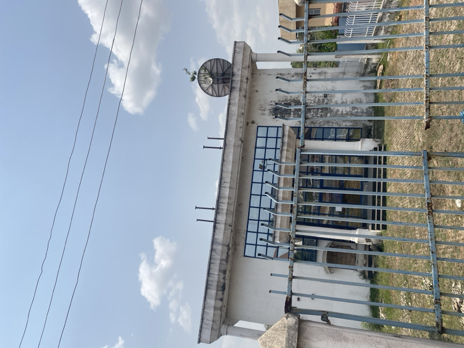 This image captures the exterior of a two-story building in Afikpo, Nigeria, observed from behind a metal fence. The primary structure is off-white with a flat roof featuring a pronounced, weathered cornice. The upper floor displays a series of large, rectangular windows with blue-tinted glass arranged in a grid pattern. Flanking the building are two classical-style columns, one partially visible on the left and another on the right. The exterior walls exhibit dirt, staining, and wear.
On the flat roof, several dark, cylindrical vents or pipes protrude vertically. To the far right of the roofline, a large, weathered, circular satellite dish is visible, with a small plant growing from its structure.
In the immediate foreground, a tall, light-blue painted metal fence with vertical bars and horizontal cross-sections stands, displaying significant rust and peeling paint. This fence partially obscures the lower section of the building and a patch of unkempt, dry grass and weeds between the fence and the building's facade.
Above the building, three parallel, dark utility wires traverse the upper portion of the frame. The sky is clear light blue with scattered white cumulus clouds. No individuals are present, and no discernible actions or interactions are occurring within the scene. To the far right, beyond the main building, another structure with a darker facade is partially visible.