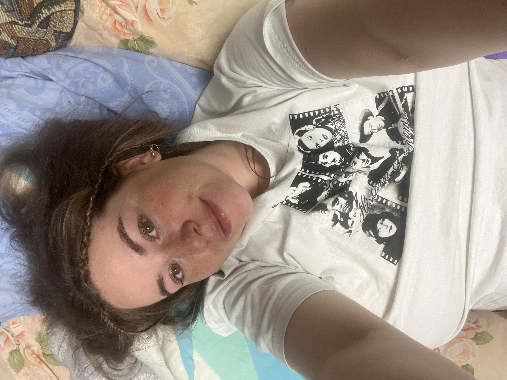 A young woman lies on a bed with patterned sheets, wearing a white t-shirt with black and white images printed on it. The location is in Chaikovsky, Russia. The woman is facing the camera with a slight smile, her hair adorned with small braids. She has fair skin with freckles visible on her face. The background includes a floral patterned pillow and bedsheets with blue and floral patterns.