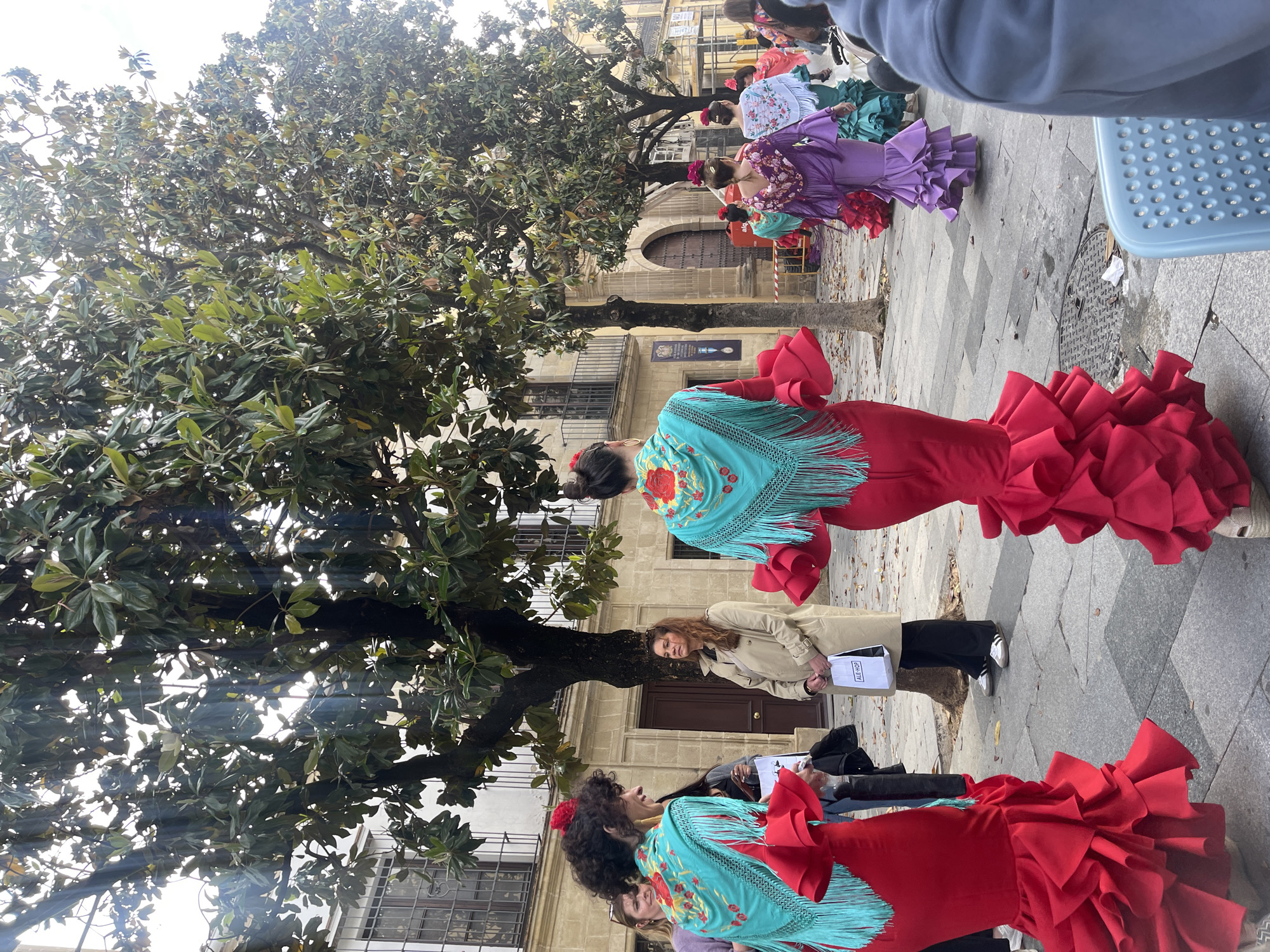 Several women are standing on a stone-paved sidewalk under a large tree in what appears to be Jerez De La Frontera, Spain. They are wearing traditional flamenco dresses in varying colors, including red and purple, with fringed shawls. Some have flowers in their hair. A woman in a tan trench coat is holding a white bag with black lettering. In the background, there is a stone building with arched doorways and windows. The scene captures a gathering or celebration in the city, showcasing its cultural traditions.