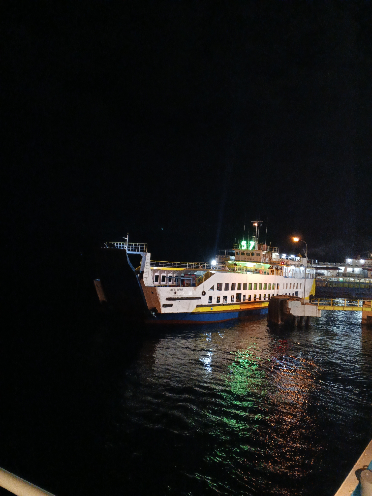 A ferry, identified by the number "B7" illuminated in green on its superstructure, is docked at a pier at night. The ferry has a white hull with blue and yellow trim. It is positioned to the left of the frame, with its stern facing the viewer. A ramp extends from the ferry to the pier, which is made of concrete supports and yellow railings. Several lights illuminate the ferry and the surrounding area, reflecting on the dark water. The sky is black, indicating nighttime. The scene appears to be a port or harbor in Melaya, Indonesia, given the location context. No people are clearly visible in the image, though faint figures may be present on the ferry's deck or the pier. The overall atmosphere is one of quiet activity at a transportation hub during the night.