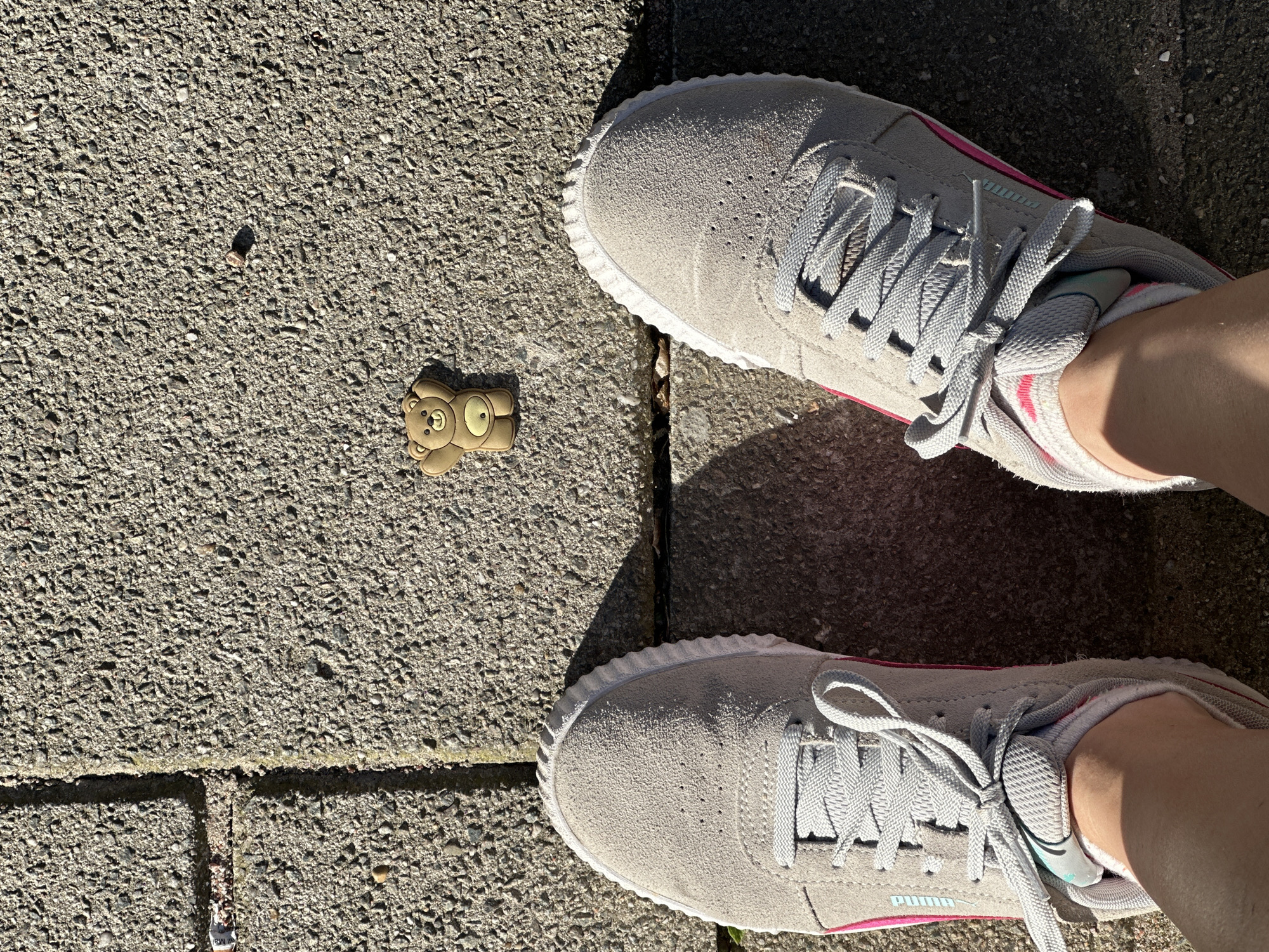 The image shows a pair of grey suede Puma sneakers on a pavement made of grey concrete blocks. A small, brown teddy bear charm lies on the pavement next to the left shoe. The background is the pavement made of the same concrete blocks. The location is not revealed in the image.