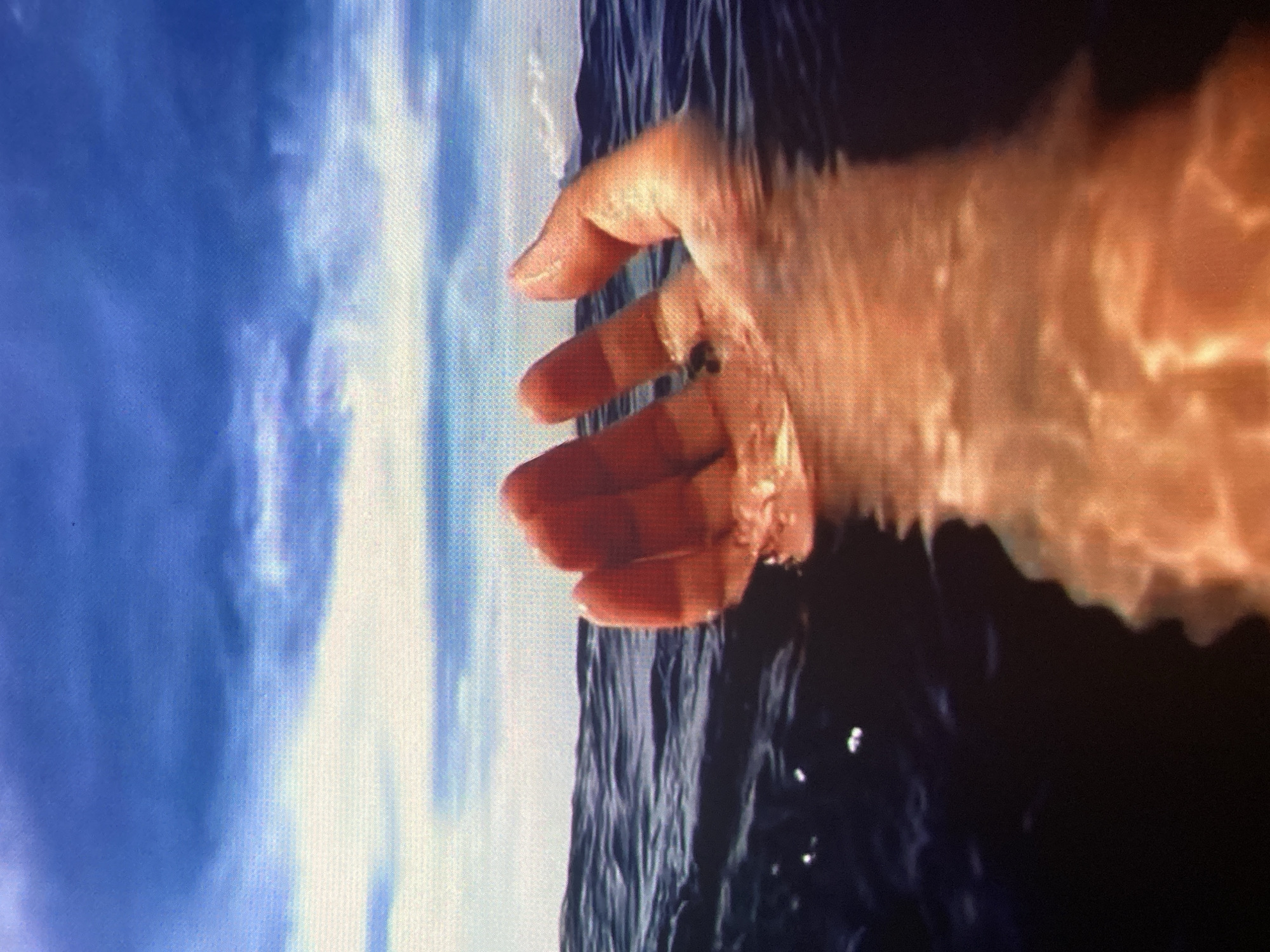 A person's hand, submerged in dark blue water, is held palm-upwards towards a bright blue sky with white clouds. The water is rippled, and the hand appears to be illuminated from above. The scene is outdoors, with the water possibly being a sea or ocean, and the sky suggesting a clear day. The image does not contain any details that would indicate it is located in the city of Ustyvytsia, Ukraine.