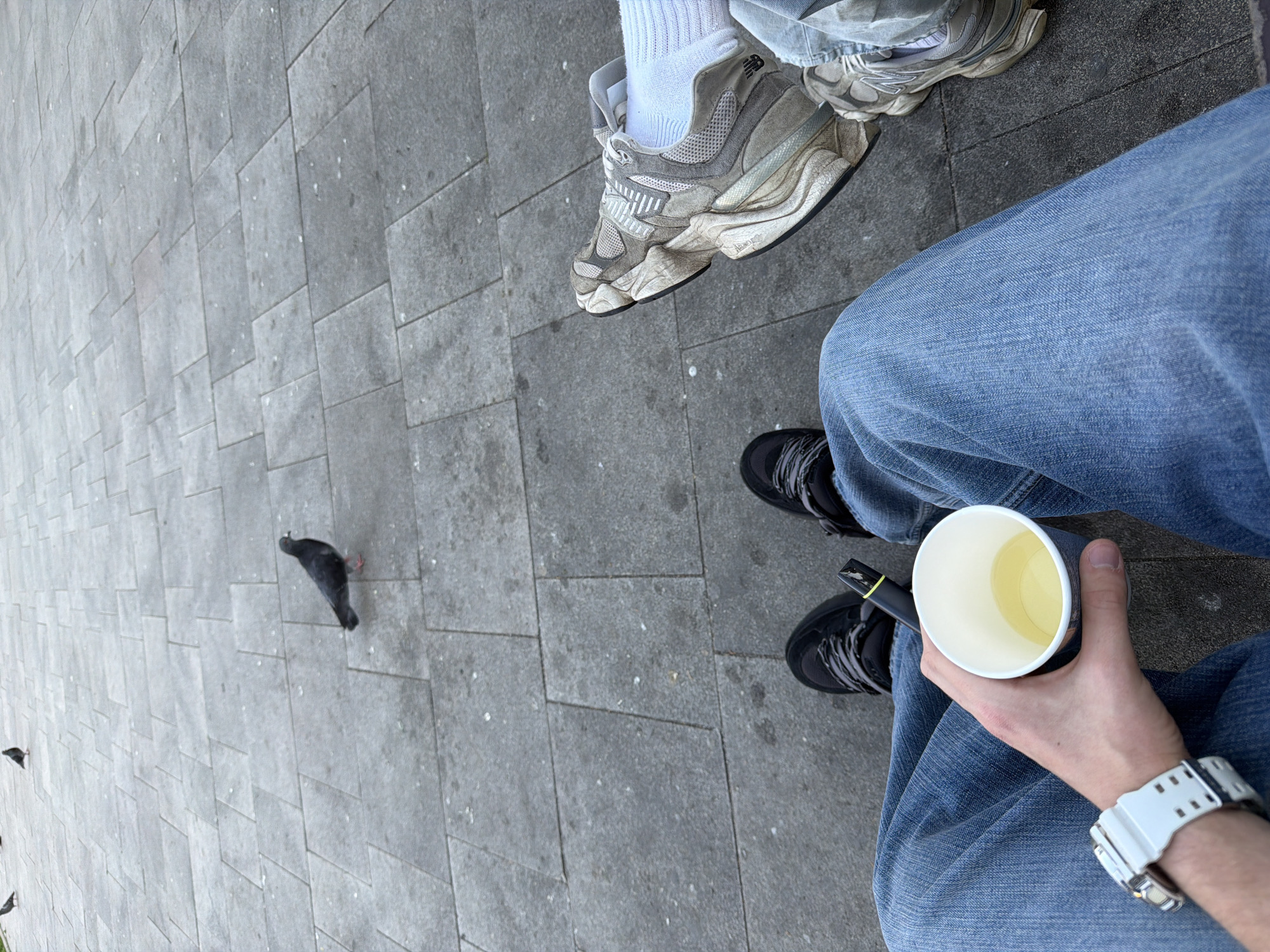 The image shows a perspective of the photographer looking down. In the foreground, a person wearing blue jeans, black shoes, and a white wristwatch holds a cup with liquid in their left hand. Visible in the upper right corner is a second person wearing an athletic shoe and white sock. On the paved surface a black pigeon walks away from the photographer. More pigeons are present at the left edge. The surface is composed of light gray brick-shaped pavers. The location is likely a city in Bulgaria, based on the user’s description.