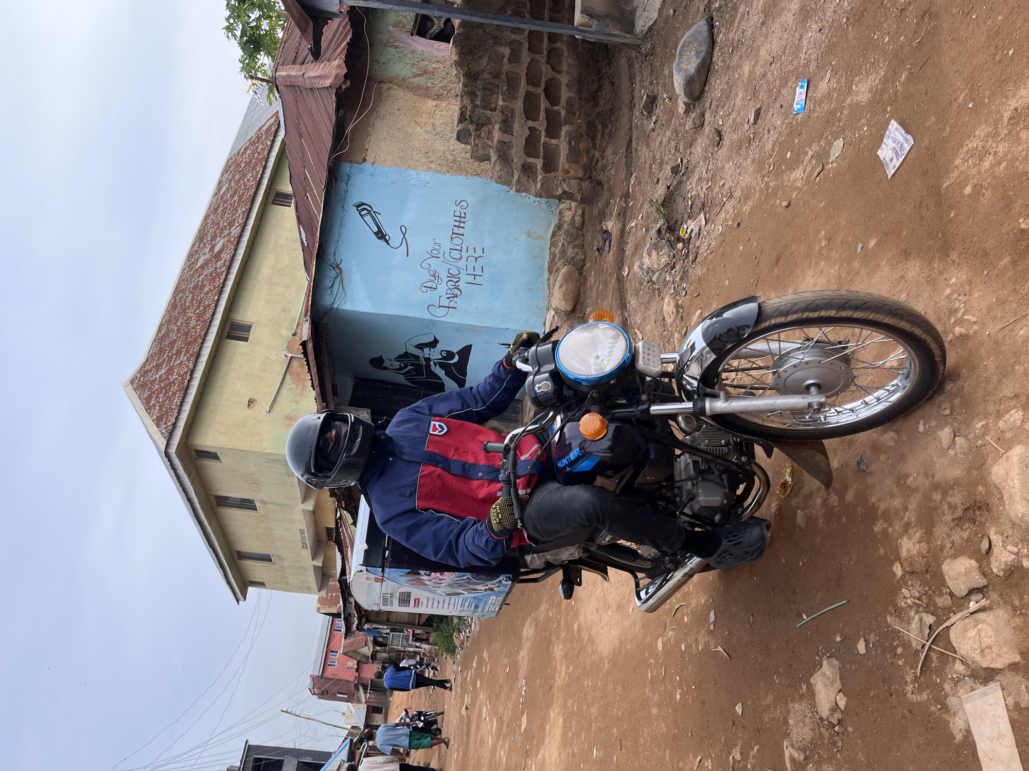 The image depicts a dirt road scene in Jos, Nigeria. A person wearing a blue and red jacket, black pants, black shoes, and a black helmet is riding a motorcycle with a large blue box attached to the back. The motorcycle has a silver exhaust pipe and a prominent front wheel with a silver rim. The background shows several buildings with corrugated metal roofs, one painted yellow and another light blue with drawings of a barber's clippers and text that reads "Dye Your FABRIC/CLOTHES HERE". There are also people walking in the background and other motorcycles. The ground is uneven and covered with dirt and rocks. The sky is overcast.