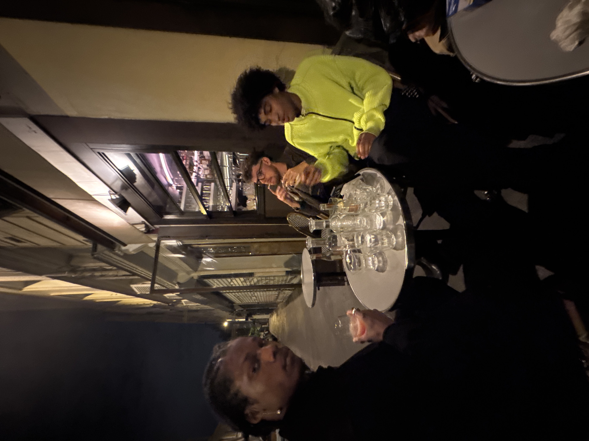 At an outdoor cafe in Paris, France, three individuals are observed at a table. One person, wearing a bright yellow jacket, is seated. Another person with glasses is next to them. A third individual, in a dark top, is in the foreground, holding a glass with a light pink liquid inside. In the background, a shelf of liquor bottles can be seen through the cafe's window. There are empty glasses on a serving platter on the table.