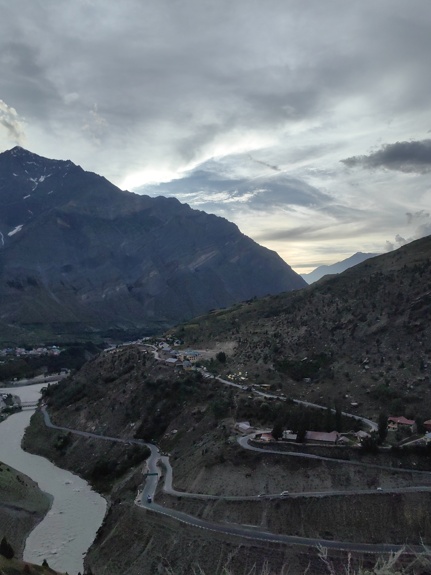 This is a high-angle, wide shot of a mountainous landscape in Gushal, India, likely taken during dusk or dawn. The sky is overcast with dramatic, wispy clouds, suggesting impending rain or recent storm activity. Dominating the scene are large, dark mountain ranges, some with snow-capped peaks visible, casting shadows over the valley below.
A wide, muddy river snakes through the valley floor, its banks lined with sparse vegetation. A winding road with multiple hairpin turns carves its way up the side of the mountain. Along this road, small clusters of buildings, possibly a village or a collection of guesthouses, are nestled into the hillside. A few vehicles are visible on the road, suggesting human activity and transit. The overall mood is serene and awe-inspiring, with the rugged natural beauty of the mountains juxtaposed with the man-made elements of the road and settlement. The lighting is subdued, characteristic of twilight, and the shadows add depth and drama to the rugged terrain. There is no visible text in the image.