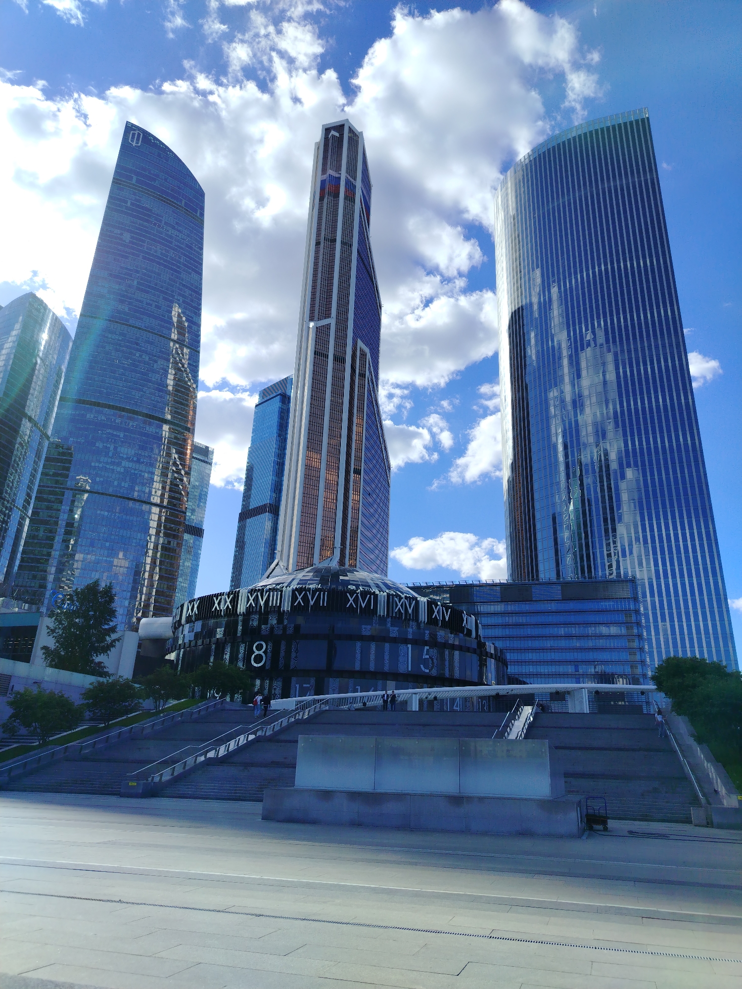 This is a low-angle shot of several modern skyscrapers against a bright blue sky with fluffy white clouds. The buildings are tall and sleek, with reflective glass facades that mirror the sky and surrounding structures. The architecture is varied, with some buildings having curved, organic shapes and others featuring sharp, geometric lines. One prominent building in the center has a multi-colored facade with red, white, and blue accents, suggesting it might be a Russian landmark.
In the foreground, there's a large, wide staircase leading up to a circular building. This circular structure has a dark, reflective exterior with large Roman numerals displayed around its circumference, resembling a clock face or a decorative element. The number "8" is prominently visible. People are seen walking on the stairs and near the circular building, suggesting it is a public space and the area is accessible.
The overall scene conveys a sense of urban grandeur and architectural achievement. The bright daylight and blue sky with scattered clouds indicate it is daytime, likely mid-morning or afternoon, with pleasant weather. The composition emphasizes the scale and height of the buildings, creating a dramatic perspective. The presence of people adds a human element to the vast architectural landscape, suggesting activity and life within this modern urban environment. The visible Roman numerals on the circular building and the overall style of the architecture point towards a location in Moscow, Russia, possibly part of a business or entertainment district.