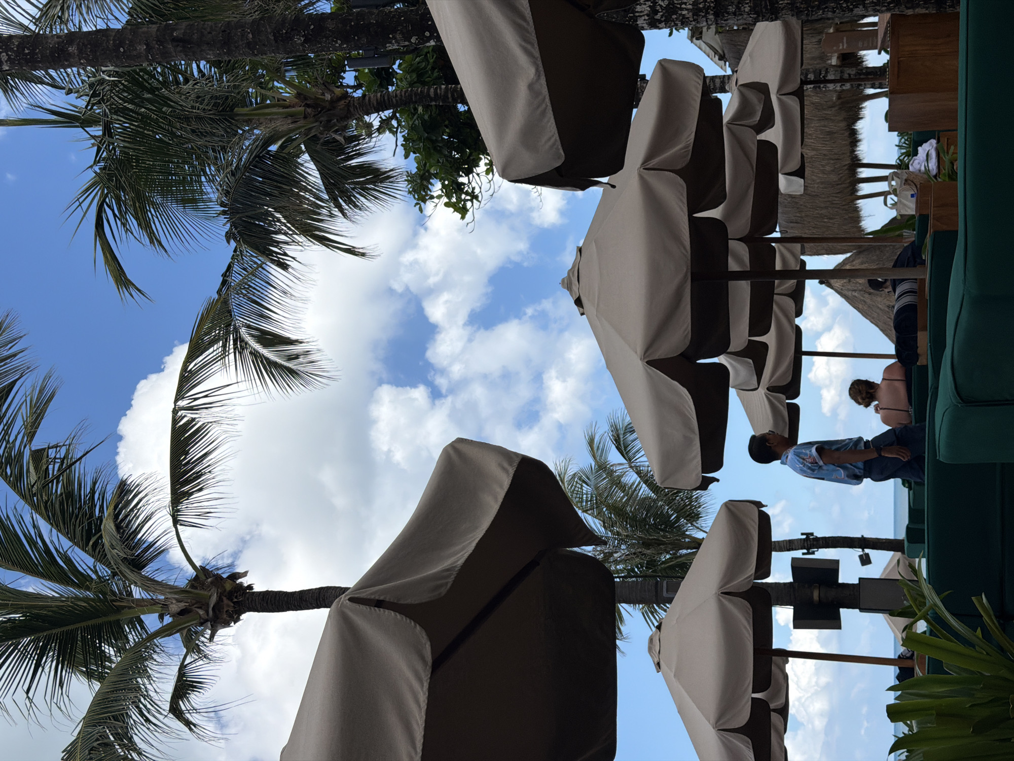 This outdoor scene in Kerobokan Kelod, Indonesia, features palm trees and several closed beach umbrellas. A blue sky with scattered clouds is visible between the trees and umbrellas. Two people are sitting on green furniture with a thatched roof in the background.