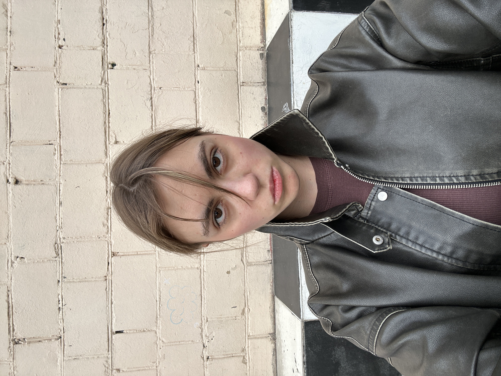 A young woman with light brown hair and fair skin is looking directly at the camera. She is wearing a dark brown leather jacket over a maroon t-shirt. Her expression is neutral, with her lips slightly pursed. Her hair is styled messily, with some strands falling across her forehead and obscuring her right eye. The background is a light beige brick wall, with some faint blue graffiti visible on the left side. Below the wall, there is a black and white checkered surface. The lighting is overcast, casting soft shadows. The scene appears to be outdoors, possibly in an urban environment given the brick wall and graffiti. No specific location in Moscow, Russia can be determined from the image.