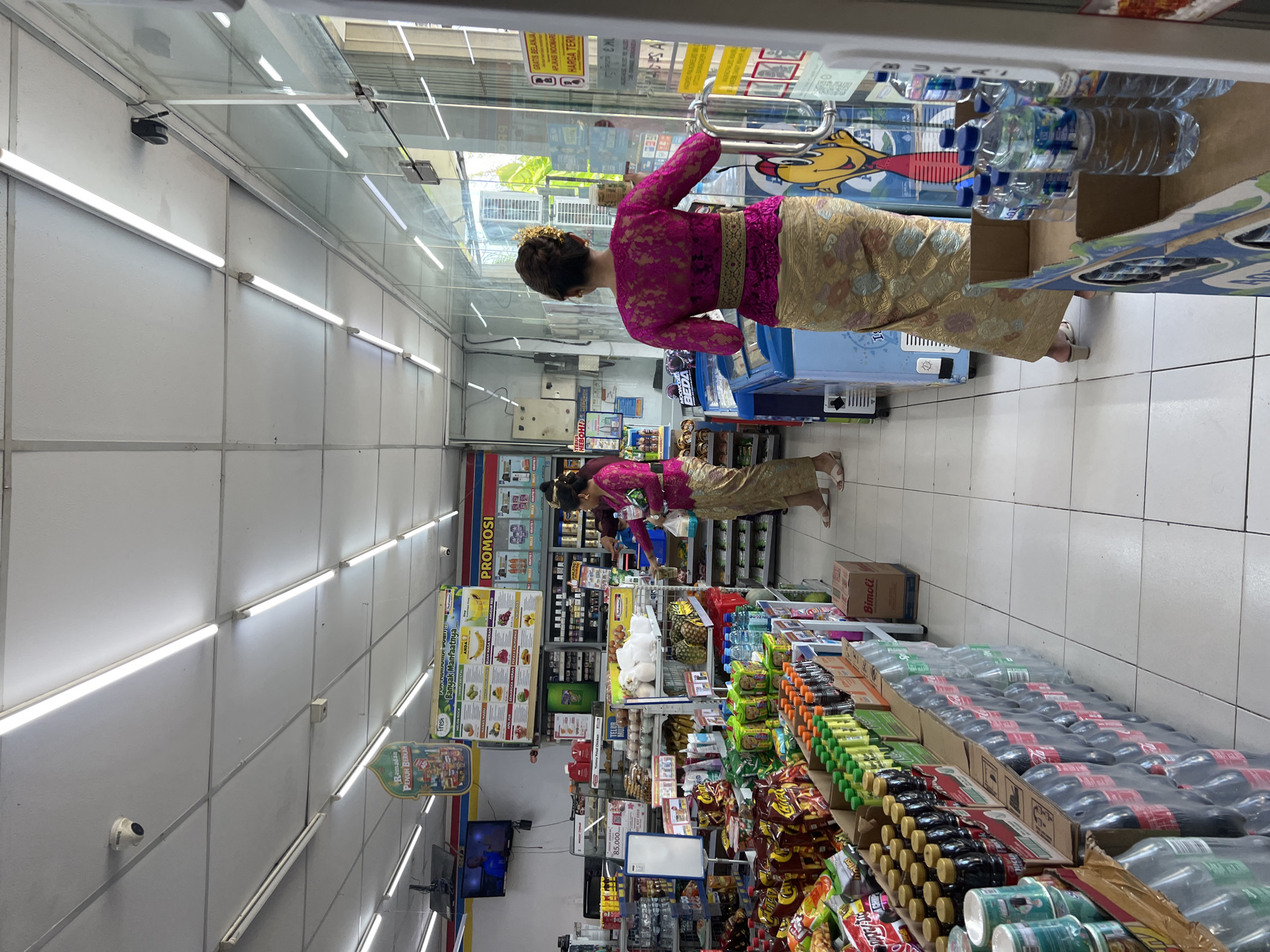 The image shows the interior of a convenience store, possibly an Indomaret, in Sukawati, Indonesia. A woman wearing traditional Balinese clothing (a pink lace top, a sarong, and a golden headdress) is reaching into a refrigerated section to grab a bottle of water. Rows of shelves are stocked with a variety of goods including bottled beverages, snacks, and other packaged items. Promotional posters hang on the shelves and walls. Security cameras are visible on the ceiling.
