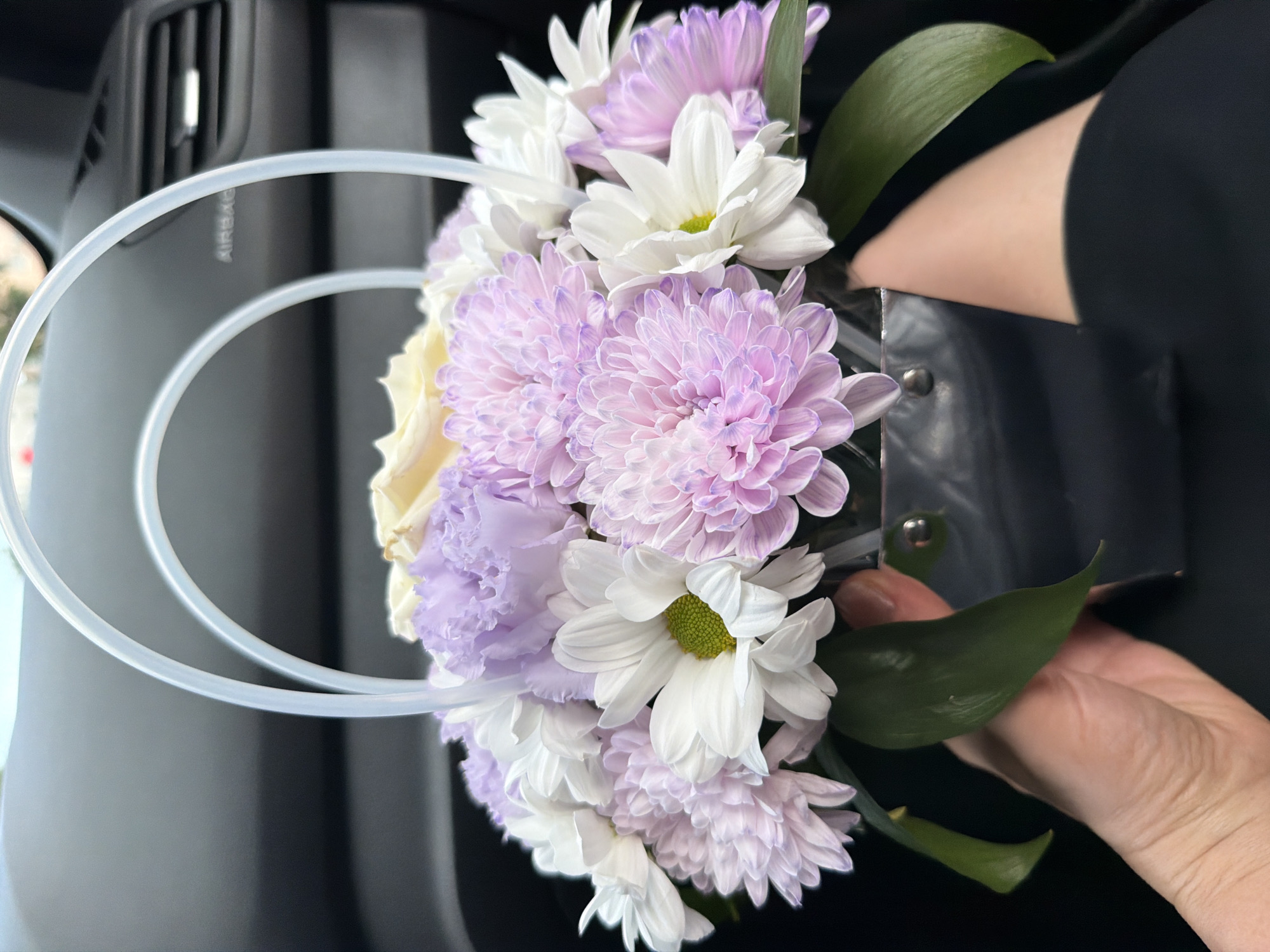The image shows a bouquet of white and lavender flowers being held in a silver container, possibly a cup holder modification. The flowers consist of chrysanthemums and other varieties. The container is being held by a hand against a black-clad arm. In the background, part of a car interior is visible, including an air vent with the word "AIRBAG" partially visible. The location of the scene is inside a car. The city is not specified.