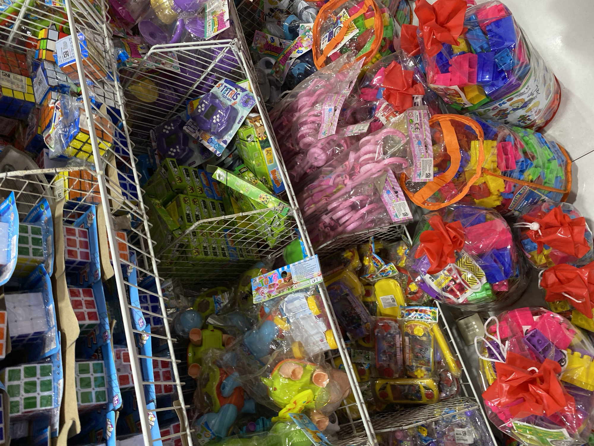 A variety of toys are displayed in a retail setting in Banda Aceh, Indonesia. The image captures several shelves filled with colorful items, including building blocks, puzzle cubes, and toy water guns. The toys are packaged in plastic bags and cardboard boxes, with some items bundled with red bows. The shelves are made of white wire metal, and the store appears to be well-stocked. The overall impression is of a toy store with a wide selection of products for children.
