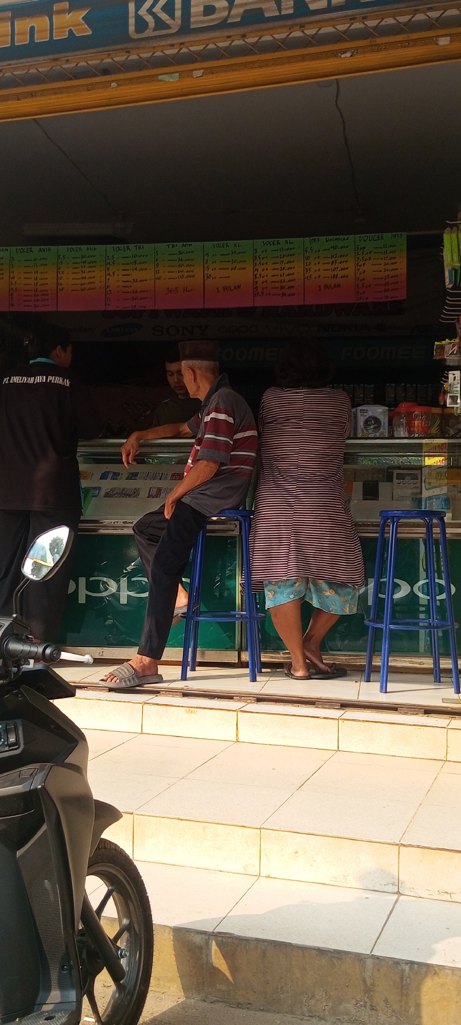 This is a medium shot, captured outdoors, of a group of people gathered around a small kiosk selling phone credit and other mobile-related services. The kiosk is brightly lit and features a price list displayed on the wall, showing various denominations and durations of phone credit. Two individuals, a man and a woman, are seated on stools in front of the counter, engaged in conversation with a third person who is partially obscured. A fourth person, standing to the left, appears to be an employee, wearing a dark shirt with a logo that reads "PT. AMELIYAH JAYA PERKAS." In the foreground, a portion of a black motorcycle is visible, suggesting a street-side location. The overall setting suggests a typical urban scene in Indonesia, likely a local marketplace or street corner in Setu. The background shows the exterior of buildings, hinting at a commercial area within the city.