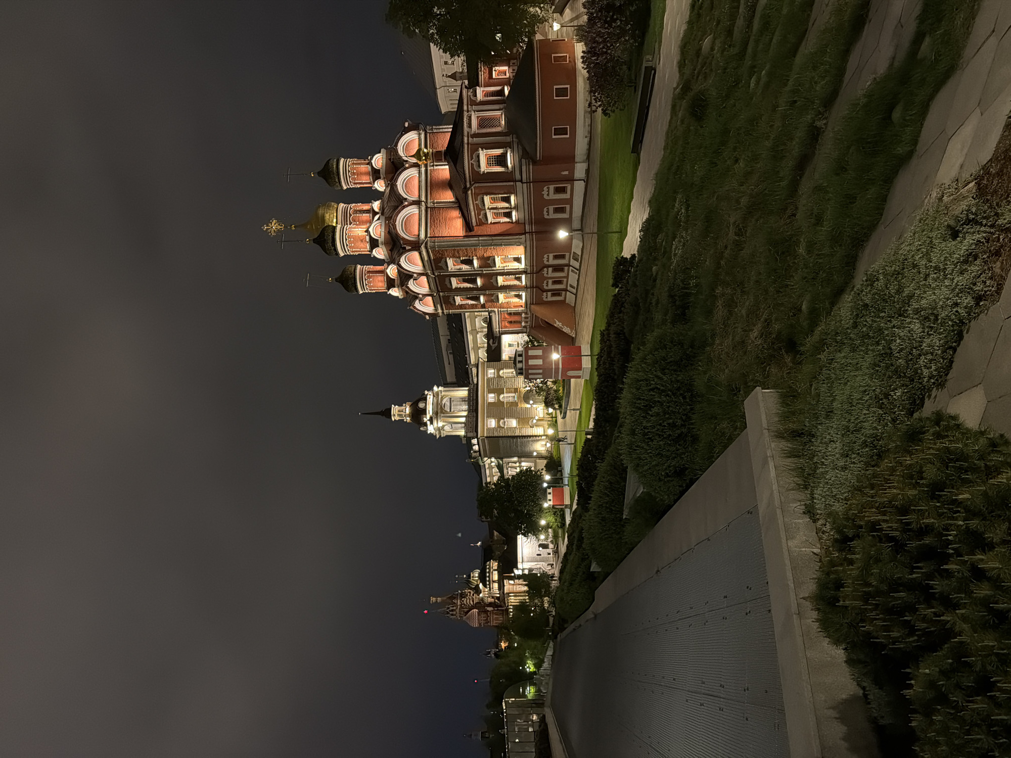This nighttime photograph shows a view of buildings and landscaping in Moscow, Russia. The architecture features traditional Russian Orthodox church structures, identifiable by their iconic onion domes, painted red and gold. These buildings appear to be lit up with warm interior lighting. A bridge or walkway runs in the foreground. The environment includes greenery such as grass and bushes alongside concrete structures and pathways. The sky appears to be overcast, providing a dark backdrop to the illuminated buildings.