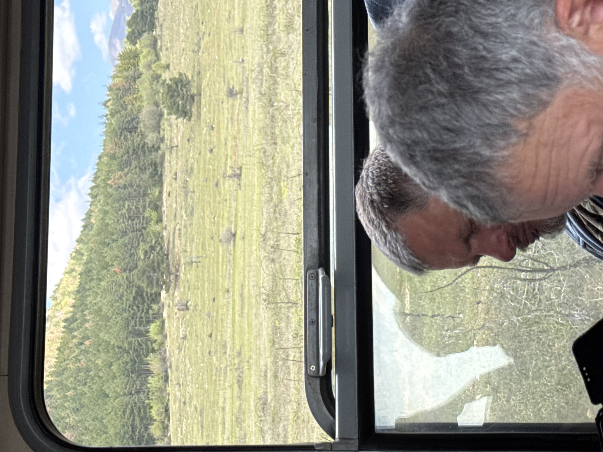 The image shows a landscape visible through a window. The landscape consists of a grassy field and a dense forest ascending a hill. Part of the window is broken. Two men are seen in the reflection of the window. The location is assumed to be Karnare, Bulgaria.