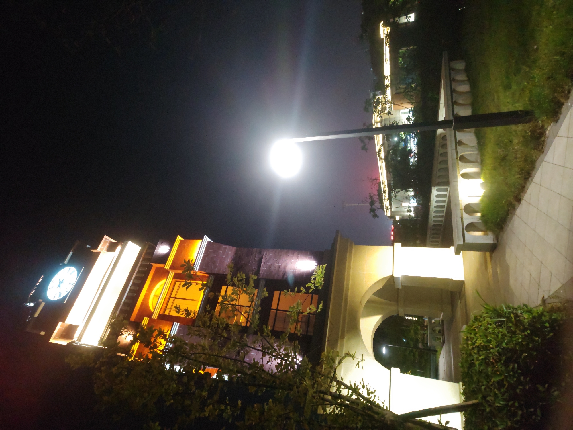 The image depicts a brightly illuminated scene at night in Chongqing High-tech Zone, China. A tall clock tower with a visible clock face is prominently featured, illuminated with white and yellow lights. The tower is partially obscured by trees in the foreground. A streetlamp with a bright circular light is positioned above the tower. A walkway with illuminated archways runs along the right side of the frame, bordered by grass and bushes. In the background, additional buildings are visible with string lights. The sky is dark.