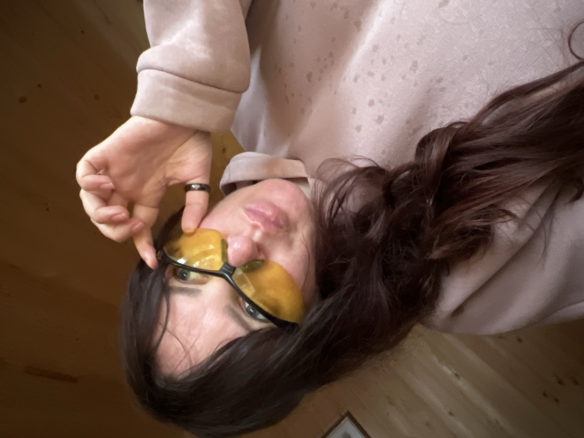 An eye-level, close-up shot captures an individual, presumably an adult female, looking directly at the viewer with her head slightly tilted. Her long, dark brown hair cascades around her. She is wearing a light beige or pale pink hooded sweatshirt. A pair of yellow-tinted glasses, possibly for night driving or blue light filtering, covers her eyes, with her right index finger touching the frame as if adjusting them. A black ring with subtle lighter speckles is visible on her right index finger. Her lips are slightly parted, suggesting a subtle expression. The background features warm-toned wooden paneling, indicating an indoor setting, possibly a room or cabin in Protasy, Russia. The lighting is soft and diffused, characteristic of indoor daytime conditions. There is no visible text in the image. The overall impression is casual and contemplative.