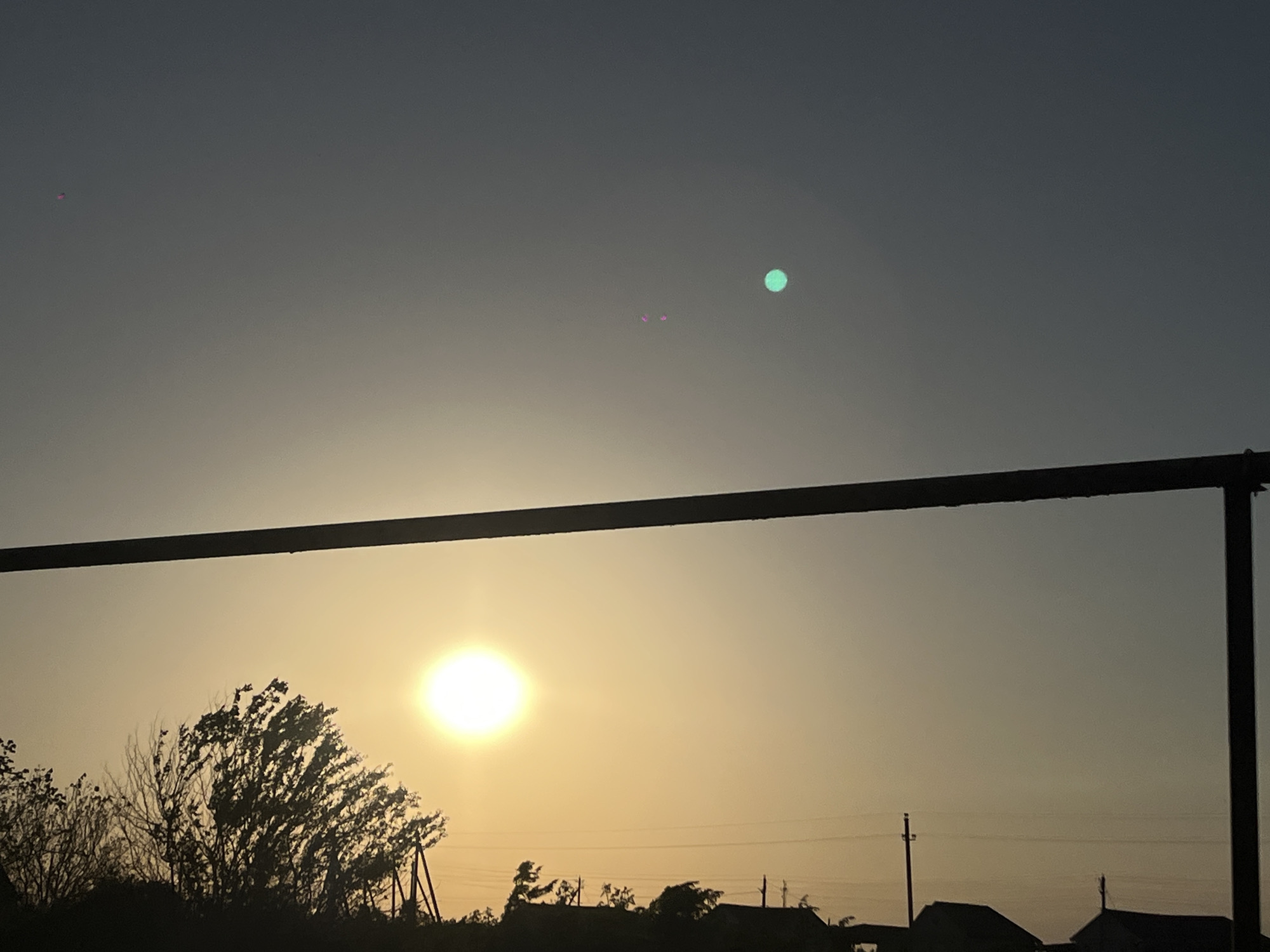 The image shows a view at dusk in what appears to be a rural or suburban area, possibly Novopokrovskaya, Russia. A dark horizontal bar and vertical post (perhaps a clothesline or part of a structure) dominate the foreground. In the middle distance, the sun is setting, casting a yellowish-orange glow behind silhouettes of trees, shrubs, and rooftops. A few utility poles are also visible. The sky transitions from light orange near the horizon to gray-blue higher up, with some lens flare artifacts visible as a green dot and some magenta spots.