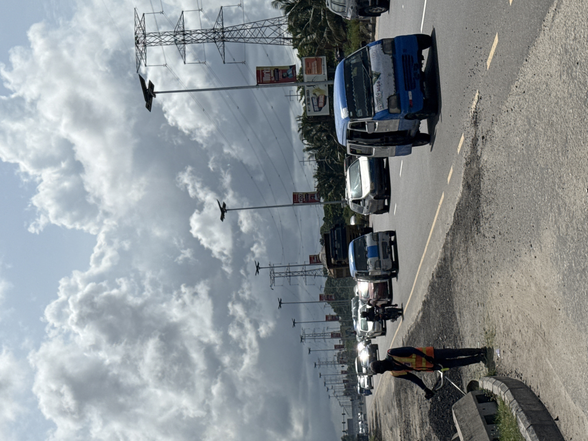 This image captures a daytime scene along a multi-lane road in Lekki, Nigeria, under an overcast sky filled with large, white clouds. The road, featuring yellow dashed lane markings, stretches into the distance on the right, with a visible dirt shoulder and concrete curb on its edge.
In the immediate foreground, a person wearing a high-visibility orange safety vest over dark attire is standing on the roadside, seemingly engaged in an activity involving a long object or tool.
A line of vehicles, including several distinctive blue and white public transport buses (typical of Lagos Danfos), cars, and a motorbike, occupies the road. Text visible on the side of one prominent blue bus includes "GET 300 CASH" and "LAGOS STATE 3054". The vehicles appear to be either in traffic or stopped.
On the left side of the road, numerous tall metal electricity pylons (transmission towers) with power lines extend far into the background, alongside several streetlights which also have small, dark, rectangular panels attached. Beneath the power lines and partially obscured by foliage, several billboards are visible. One prominent billboard shows "iWORK" on an orange background, and another smaller sign mentions "200% DATA". The background also features lush green vegetation, including palm trees, bordering the road. The scene suggests a busy urban or semi-urban environment.